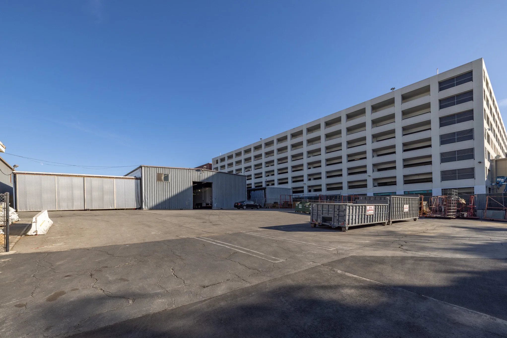 Empty parking lot with a large multi-story white building, metal containers, and construction equipment under a clear blue sky.