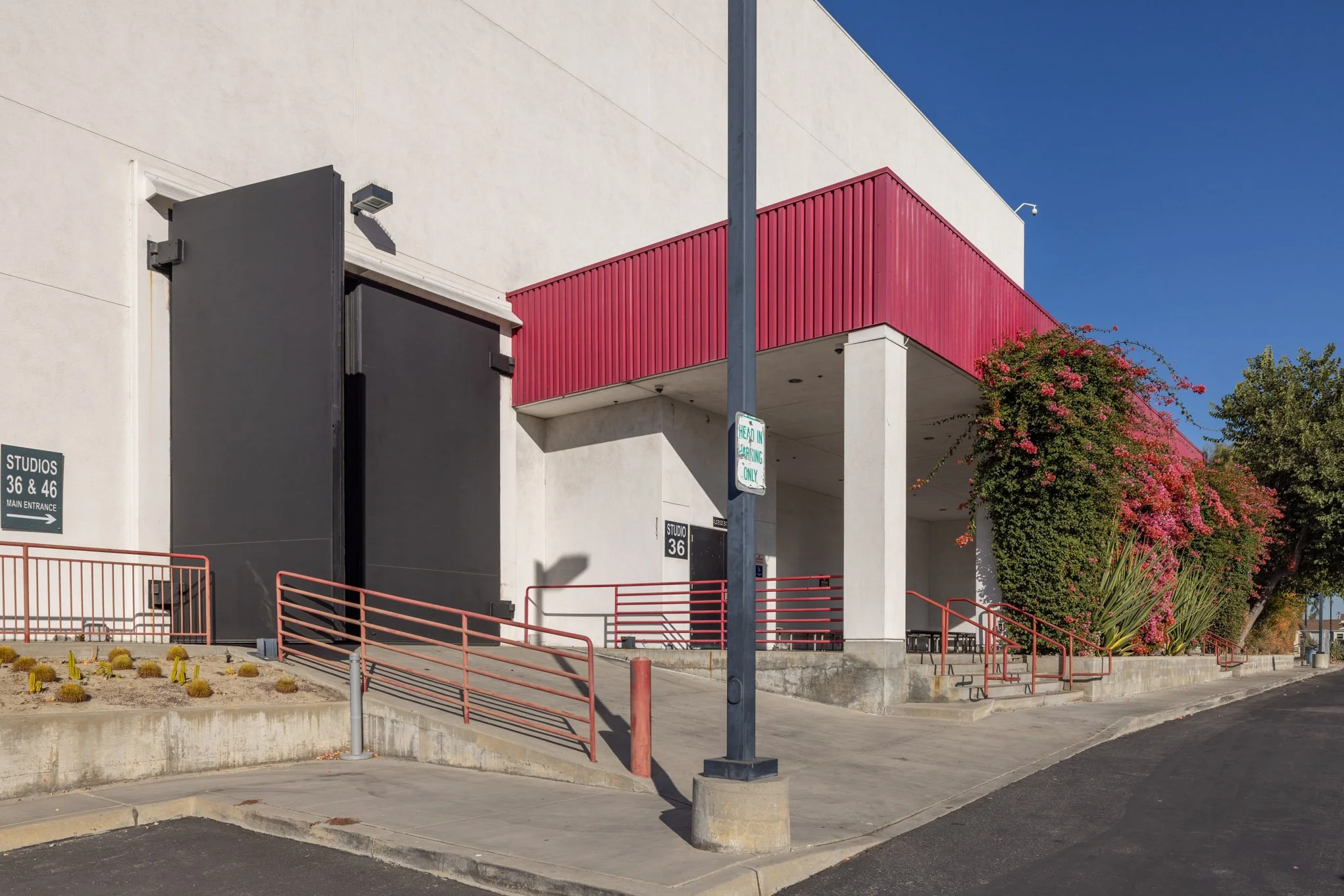 Exterior of a modern building with graffiti-covered black door, white walls, red awning, stairs, and pink flowering bushes, with a street and blue sky.