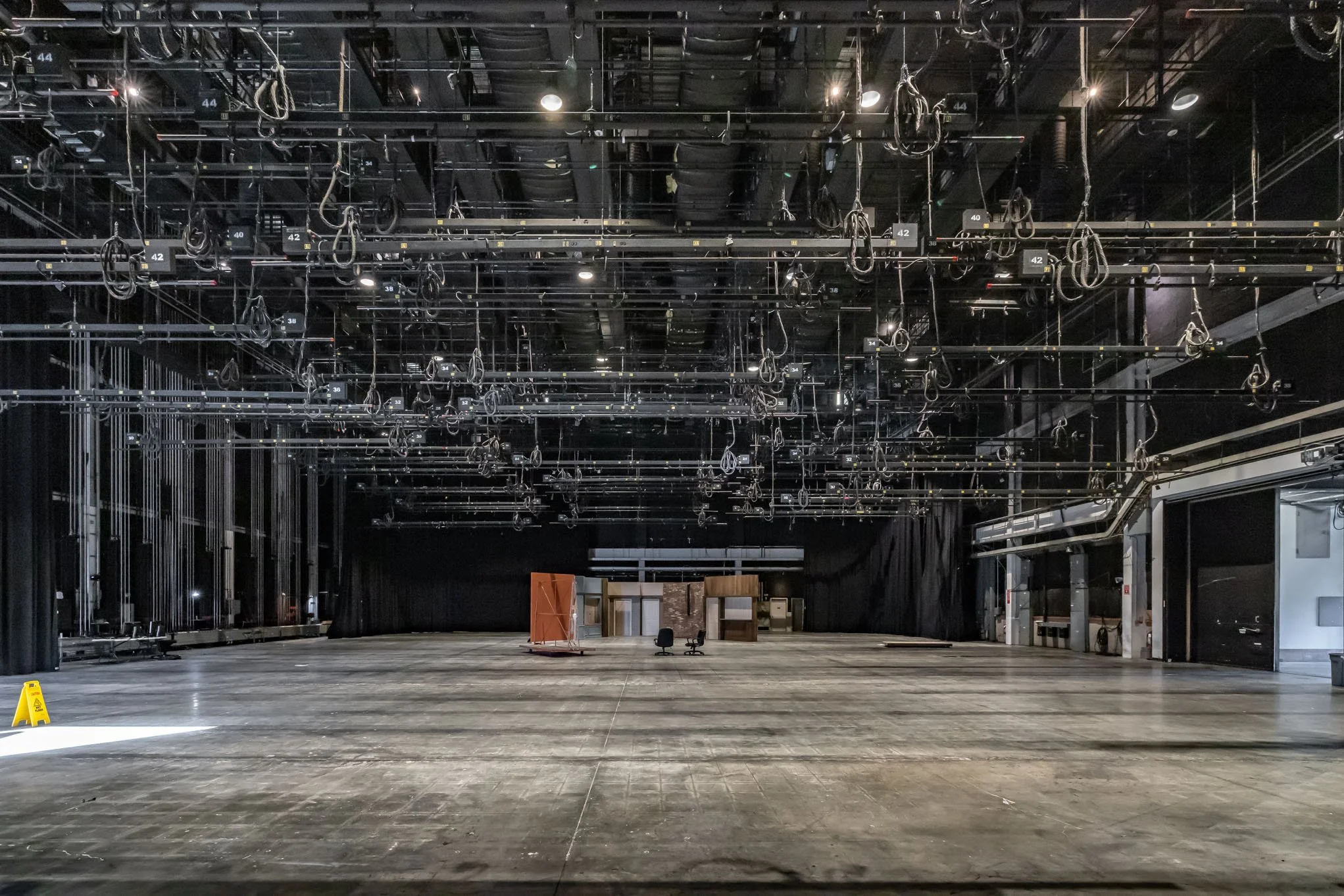 Empty theater stage with black curtains, lighting and rigging on ceiling, and some set pieces at the back, including a small structure and chairs.