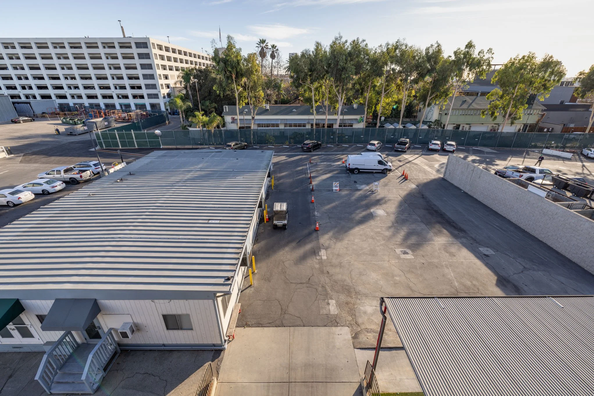 Empty parking lot with a white van and orange traffic cones, surrounded by buildings and trees.