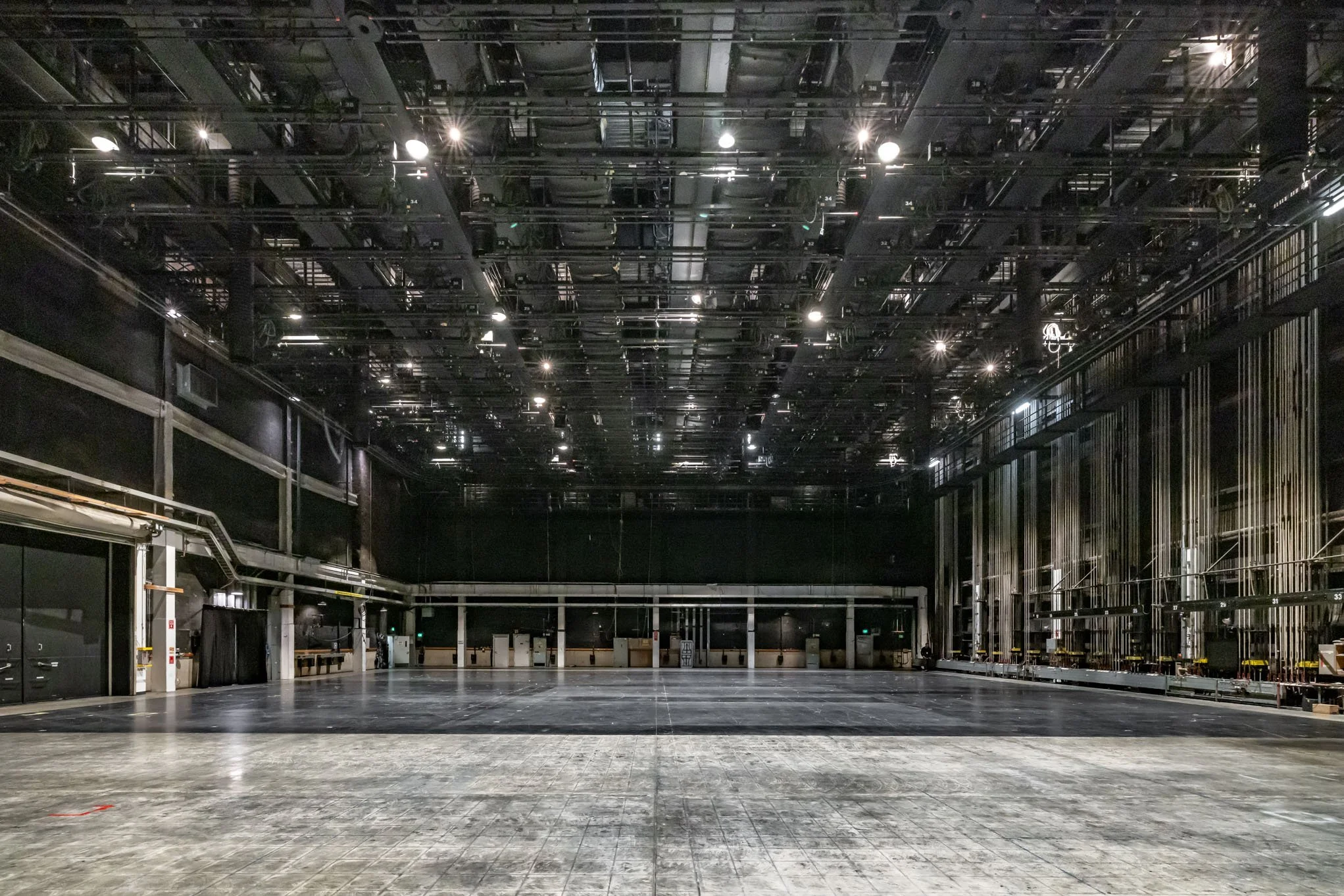 Empty stage and backstage area of a theater or performance venue with black walls, overhead lighting, and rigging for curtains and scenery.
