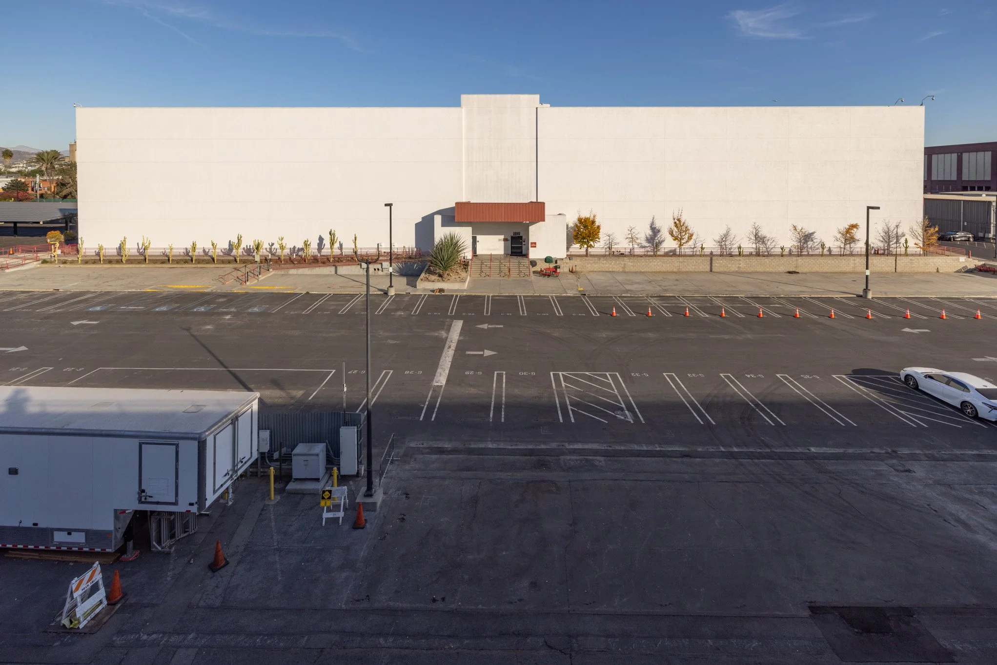 Empty parking lot in front of a large white building with small trees and desert plants along the sidewalk, and a few orange cones near the entrance.