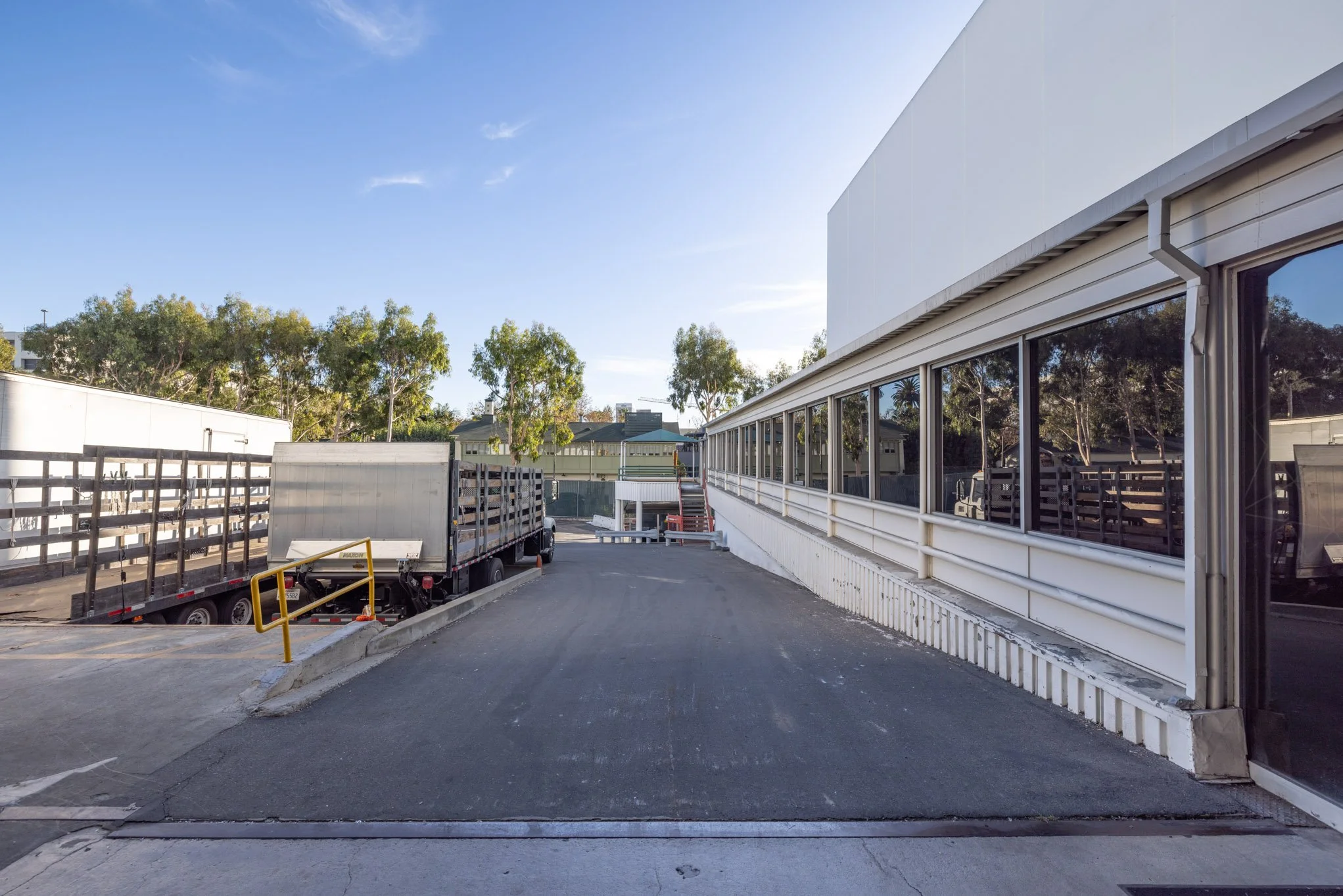 Exterior view of an industrial building with large glass windows reflecting trees, parked trucks with open trailers, and a clear blue sky.