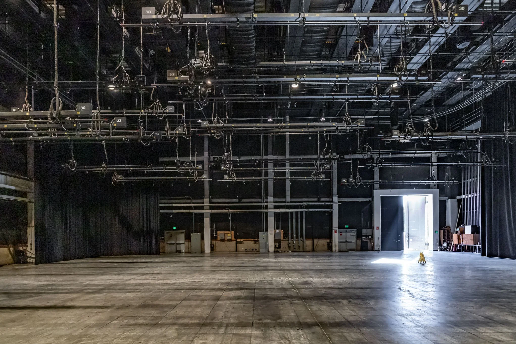 Empty stage with black curtains, overhead stage rigging, and a bright open door at the right, letting in natural light.