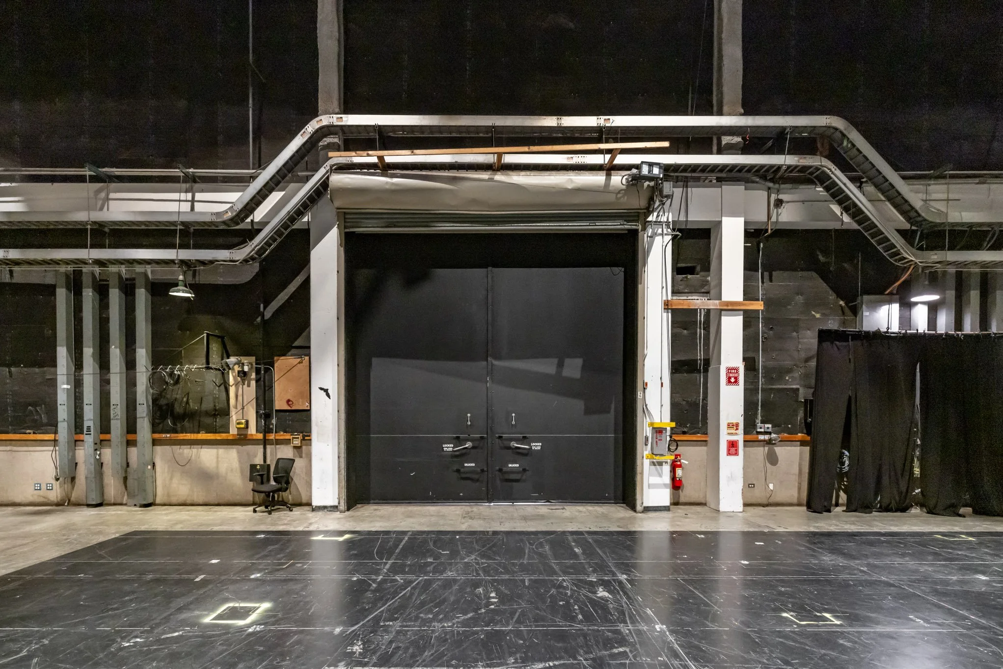 Empty stage with black curtains, chairs, and equipment in a theater or performance space.