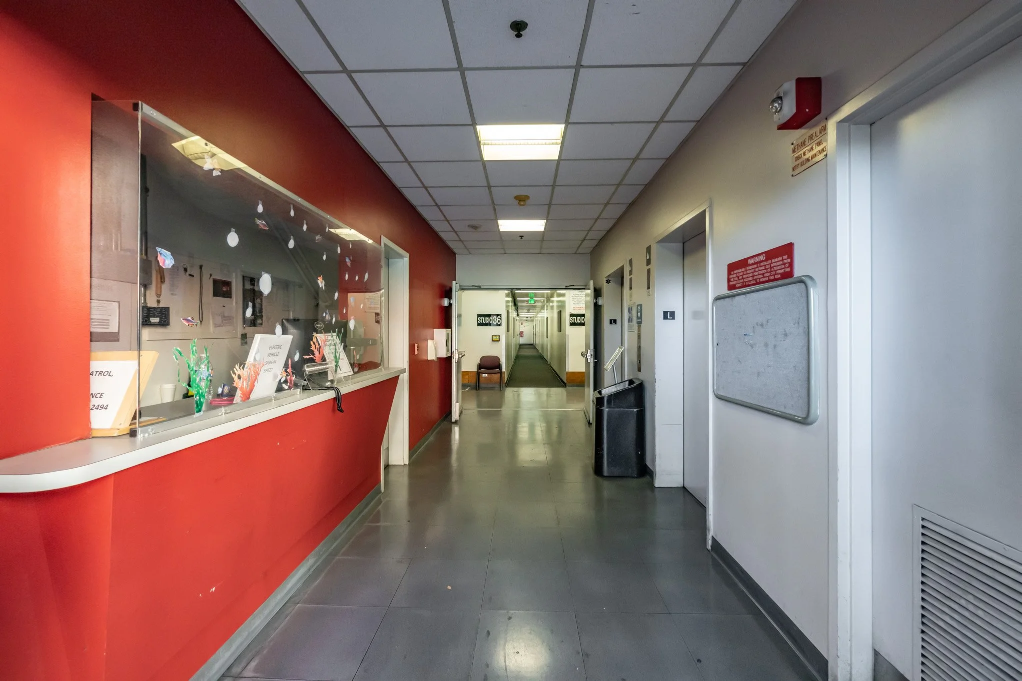 Inside a building hallway with a red reception window on the left and elevator doors on the right. The hallway extends into the distance with signs labeled 'STUDIO 36'.