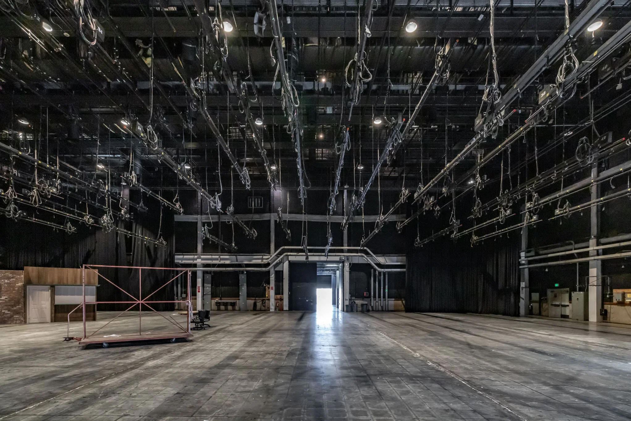 Empty theater stage with lighting, stage rigging equipment hanging from ceiling, and an open back entrance with bright light