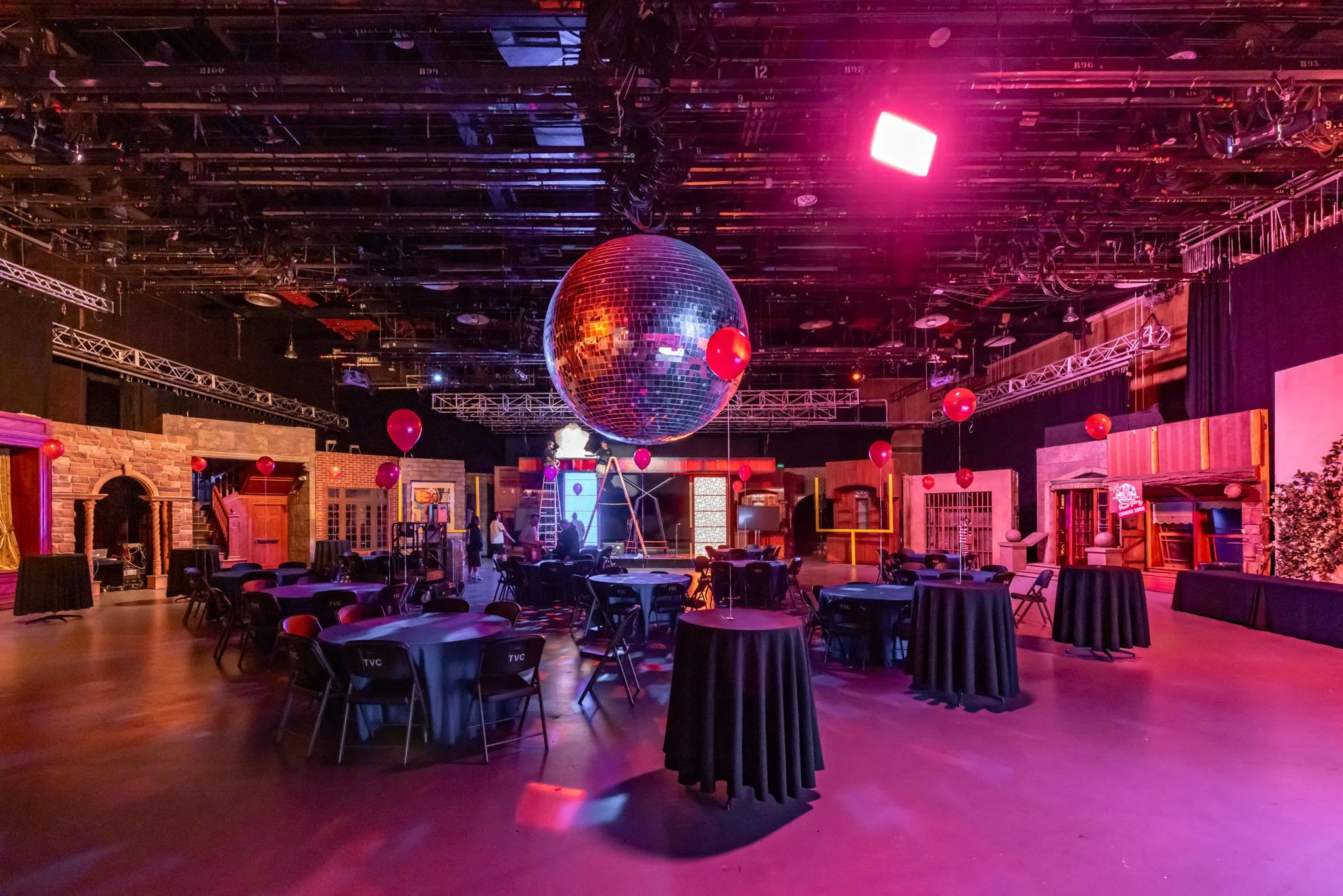An empty indoor event space with round tables covered in black tablecloths, black chairs, and decorative red balloons hanging from the ceiling. There is a large disco ball in the center, with stage lighting creating colorful reflections. The backgrou