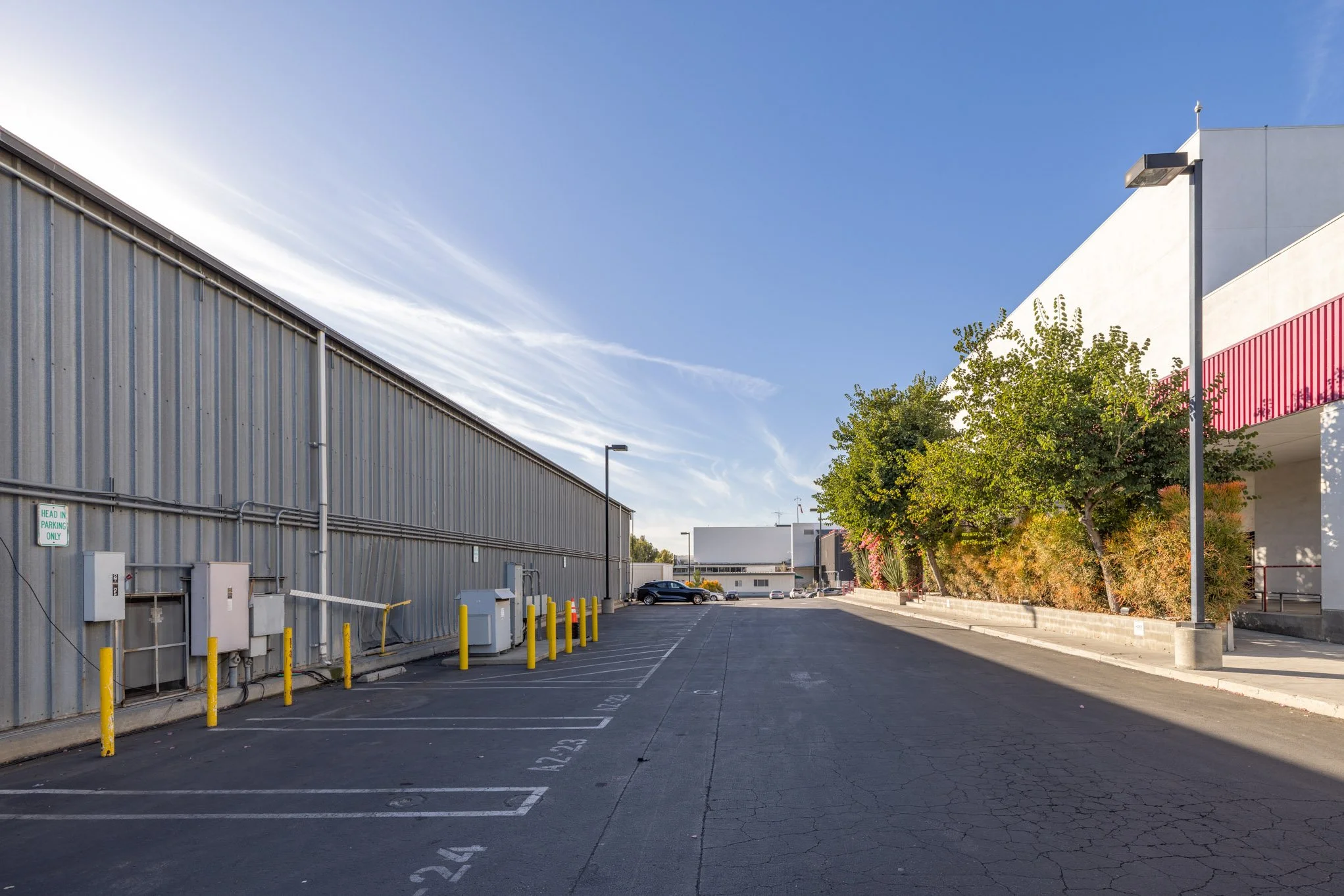 Empty parking lot next to a large industrial building with a metal exterior on the left and a modern white building with red accents on the right. There are a few cars in the distance, trees, and streetlights under a clear blue sky.