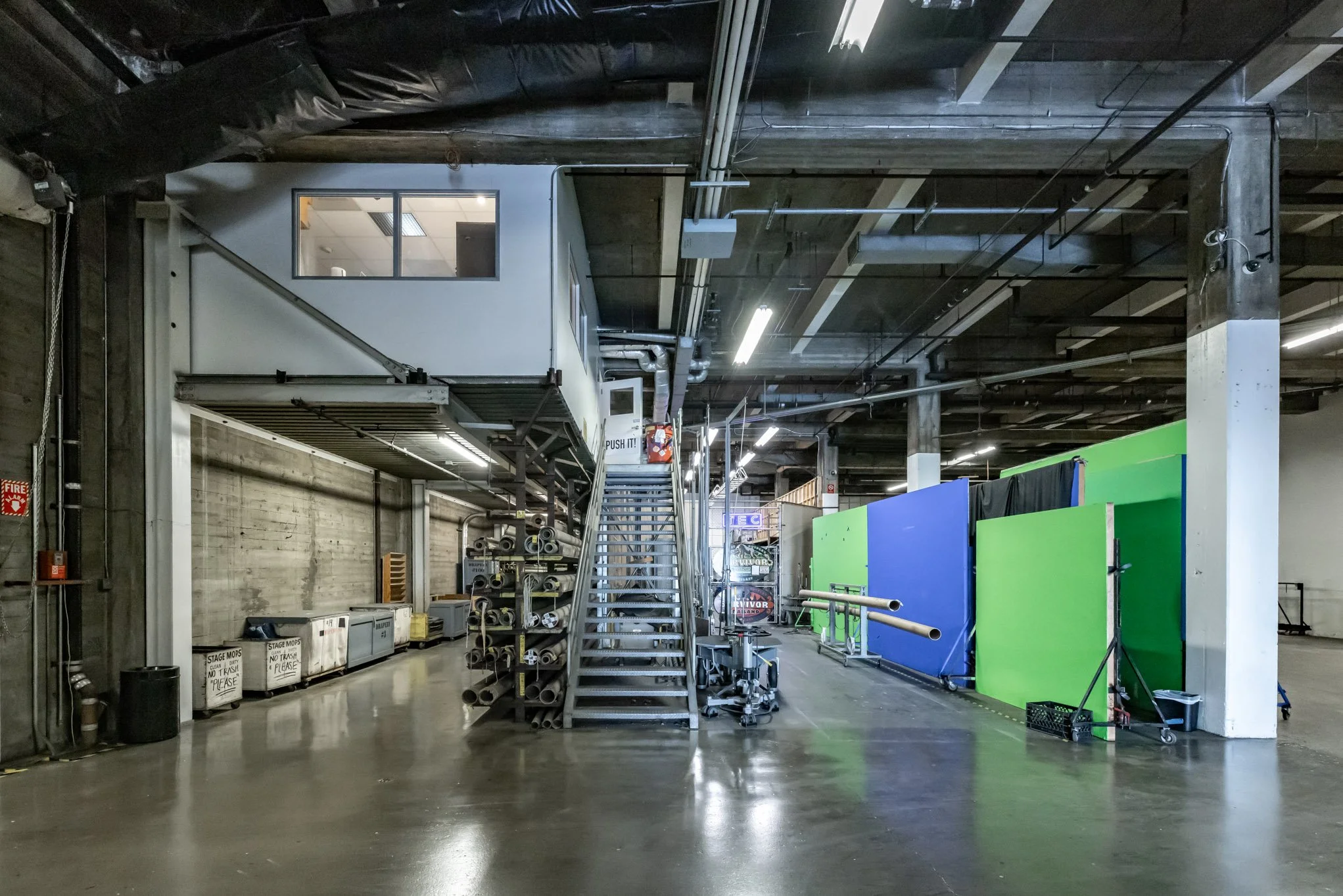 Industrial warehouse space with metal staircase, storage bins, colorful partition walls, and pipes on the ceiling.