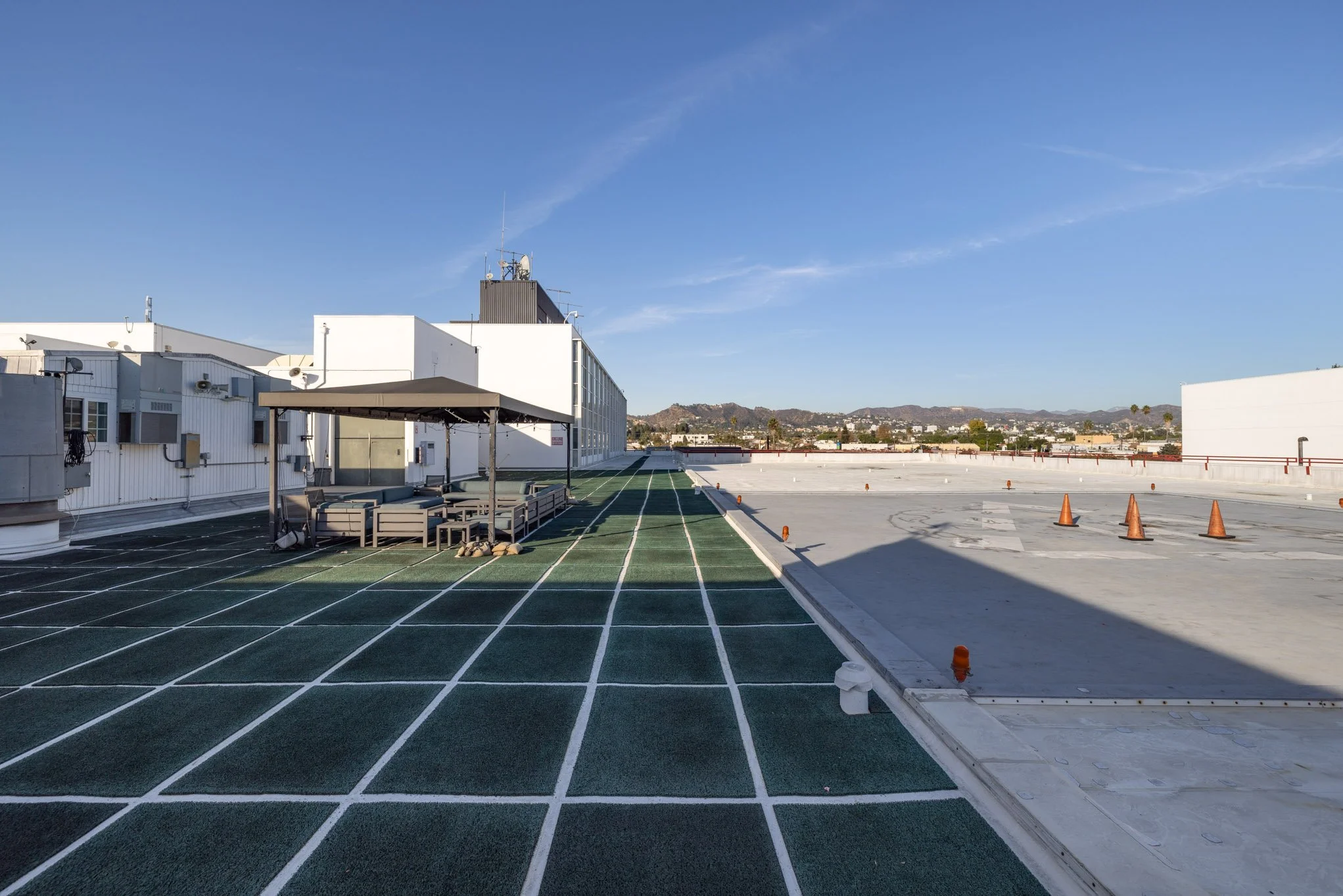 Empty rooftop with green running track on the left, large white building and equipment on the right, orange traffic cones in the background, and mountains in the far distance under a clear blue sky.
