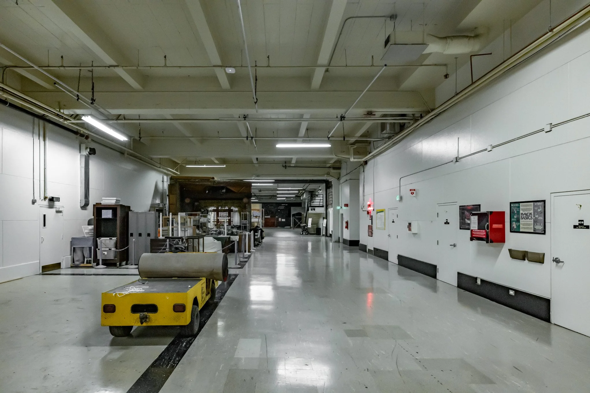 Empty indoor hallway with scattered equipment and a small yellow cart on the floor, white walls, and fluorescent ceiling lights.
