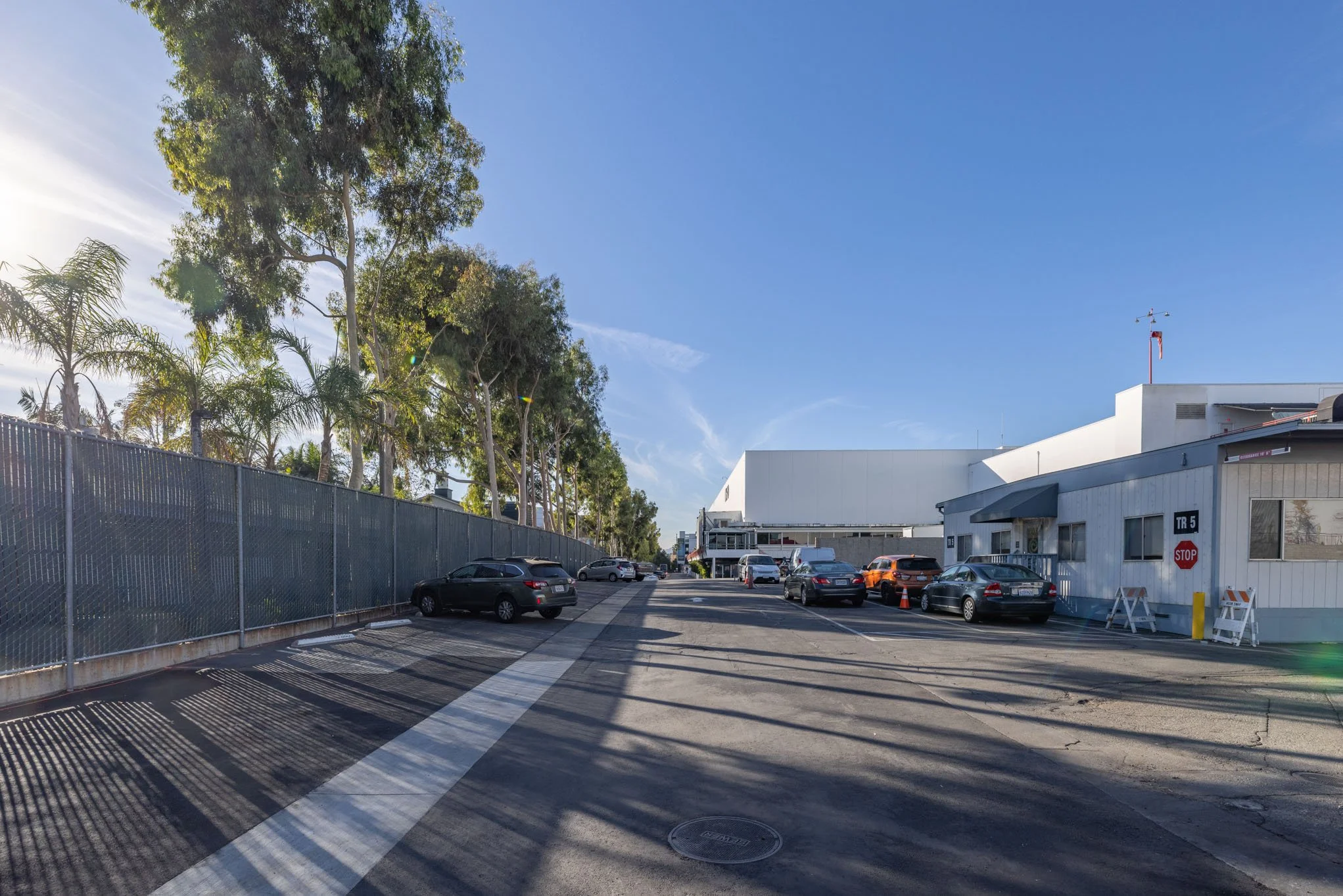 Parking lot with several parked cars, a white building on the right, a fence on the left, and tall trees behind the fence under a clear blue sky.