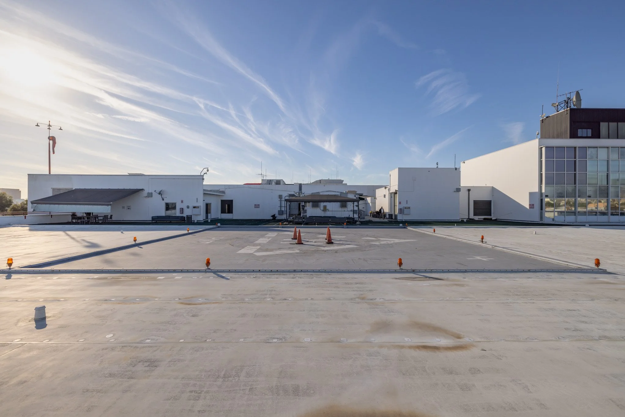 View of a building rooftop parking deck with white buildings, orange safety cones, and a blue sky with clouds.