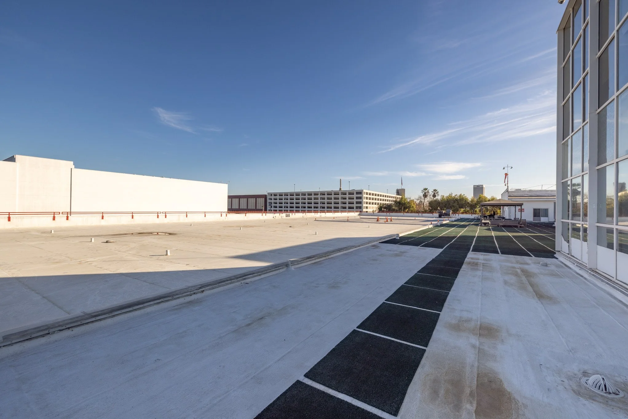 Empty rooftop parking area with marked parking spaces, a small building with outdoor seating, and a city skyline with tall buildings in the background under a clear blue sky.