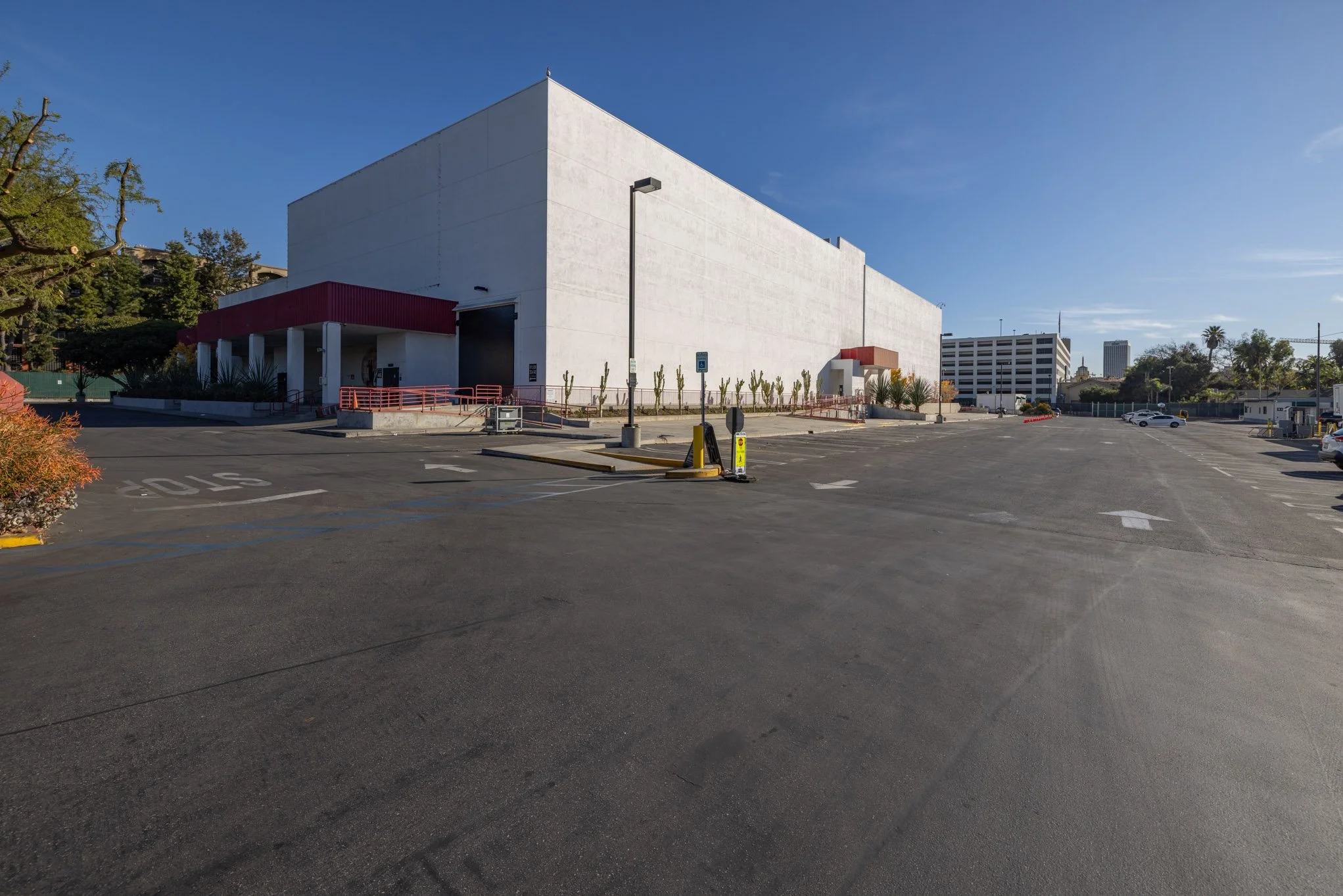 Empty parking lot in front of a large, white building with a red entrance canopy, surrounded by a few trees and some tall buildings in the distance under a clear blue sky.