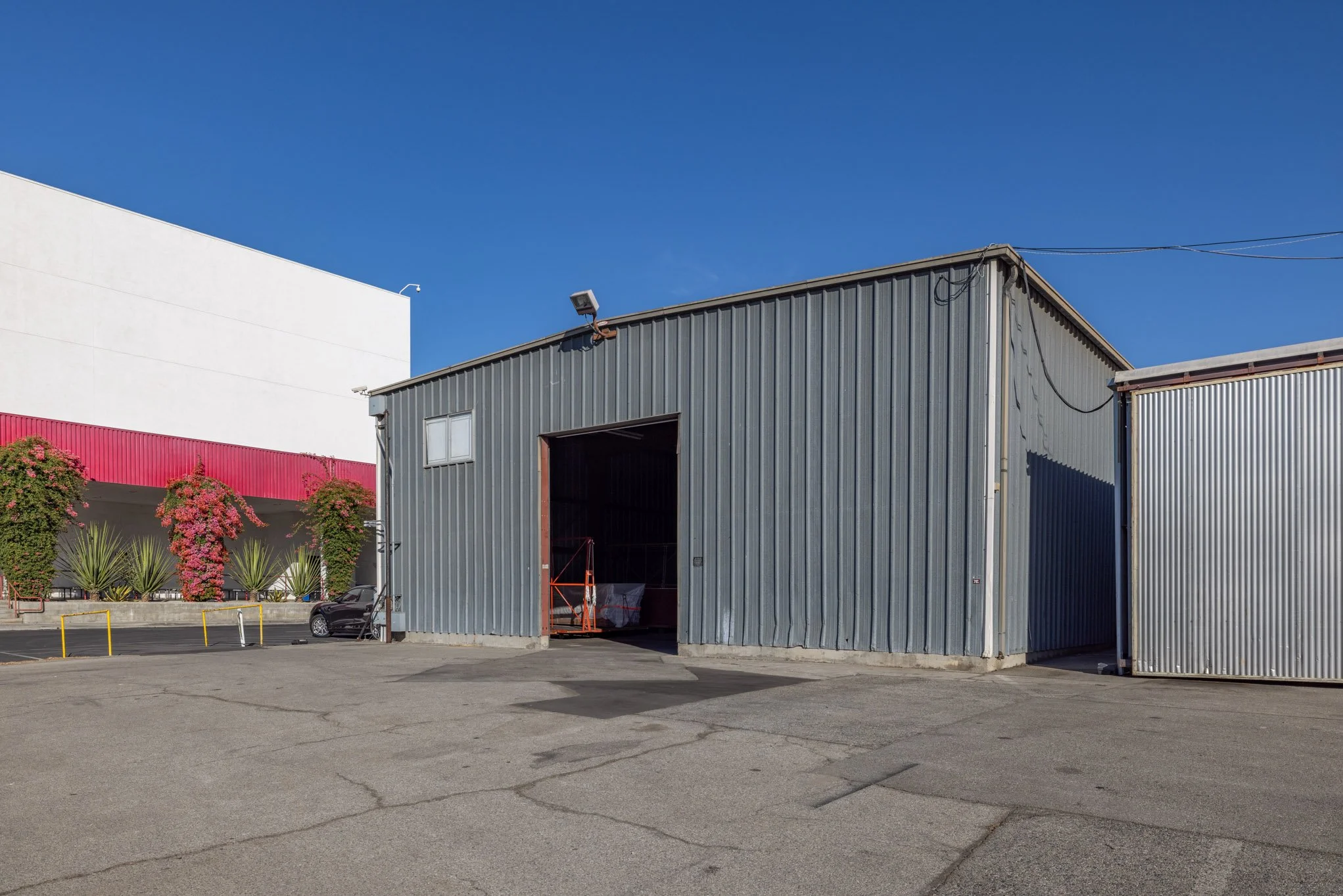 A large gray metal warehouse with an open garage door, parked car, and surrounding plants under a clear blue sky.