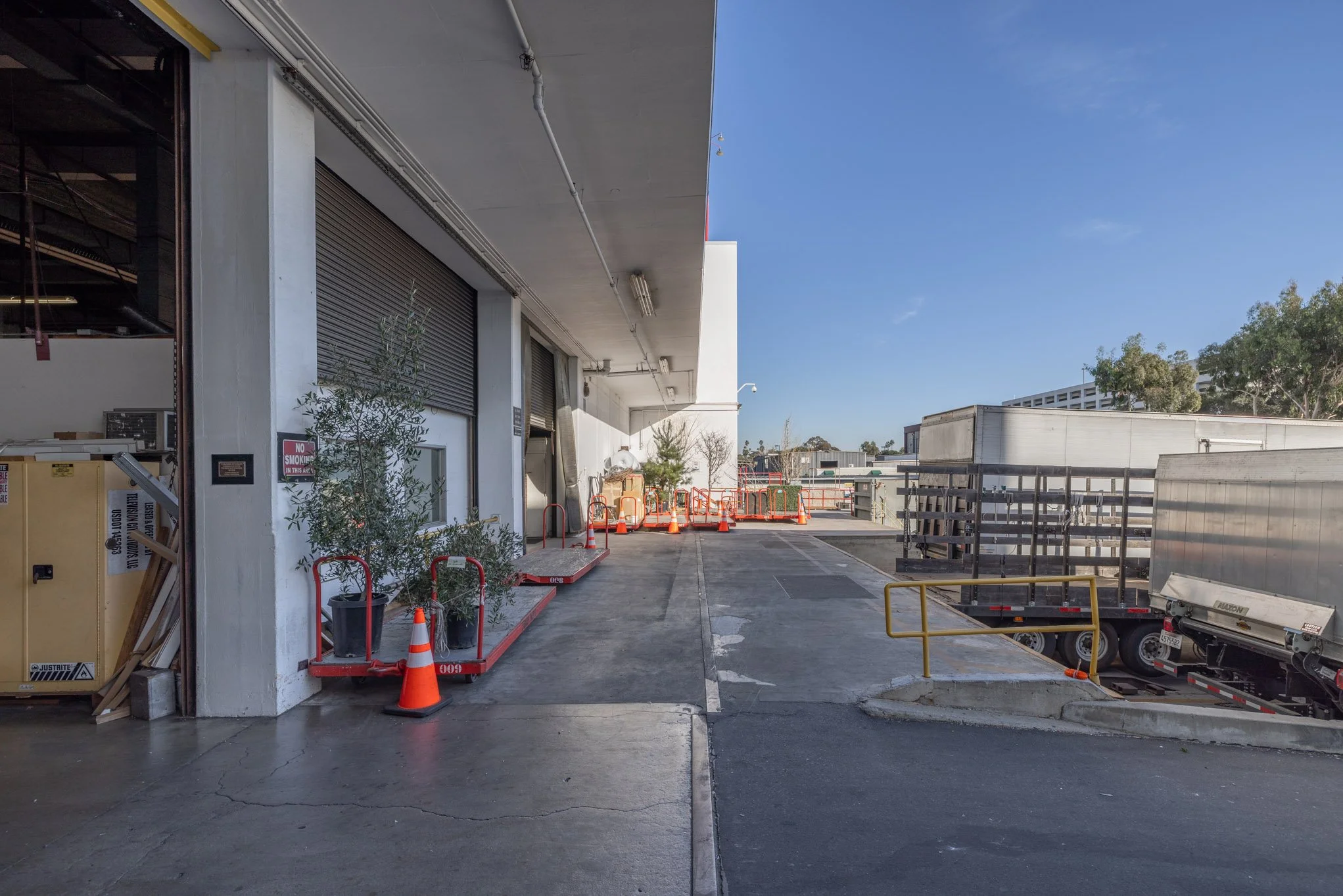 Loading dock area with orange and white safety cones, plants, a partially open roll-up door, and a long trailer truck parked on the right side, under a clear blue sky.