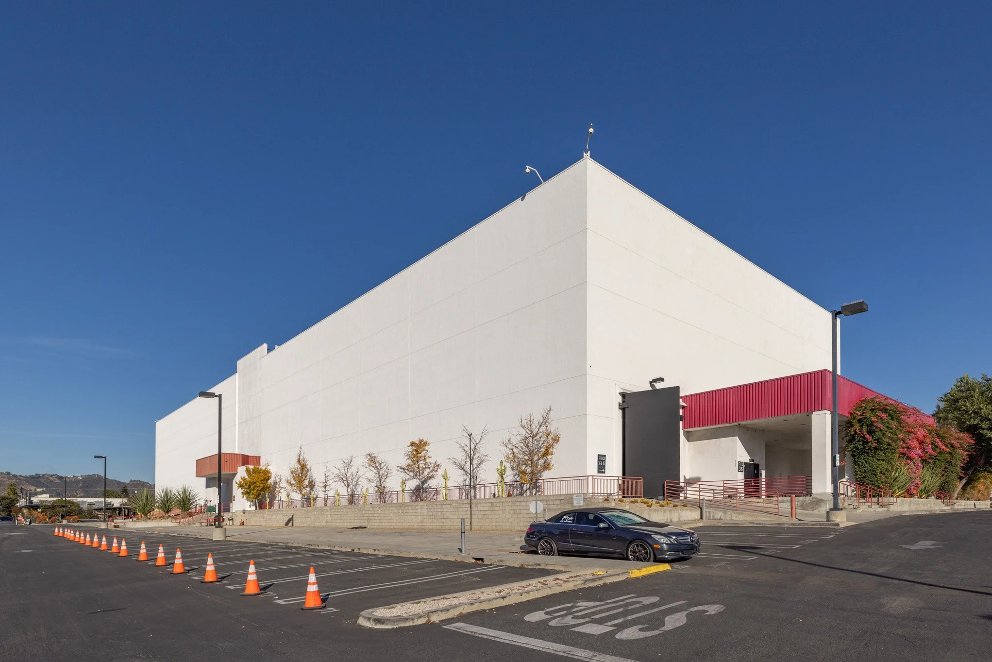 Large white commercial building with minimal windows, red accents on the entrances, and a parking lot in front with a few parked cars and traffic cones, under a clear blue sky.