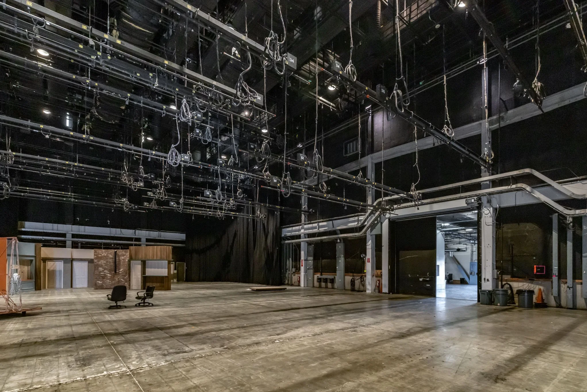 Empty theater or performance venue stage with lighting and sound equipment hanging from the ceiling, black curtains, a few chairs, and a door leading backstage.