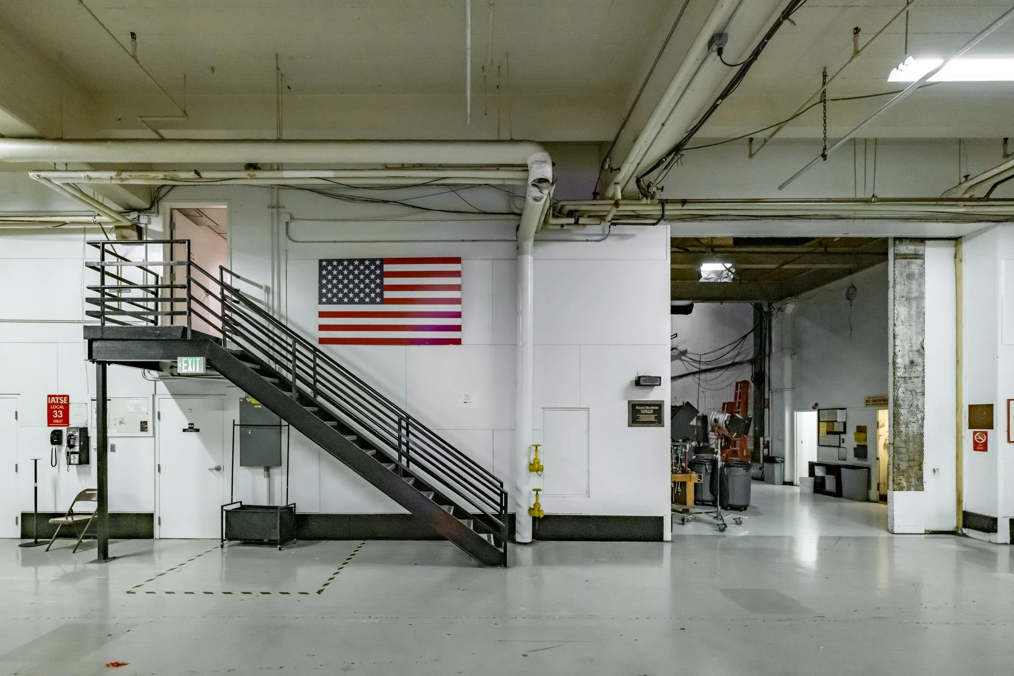Interior of a building with an American flag on the wall, a staircase, fire safety equipment, and a partially visible workshop area in the background.