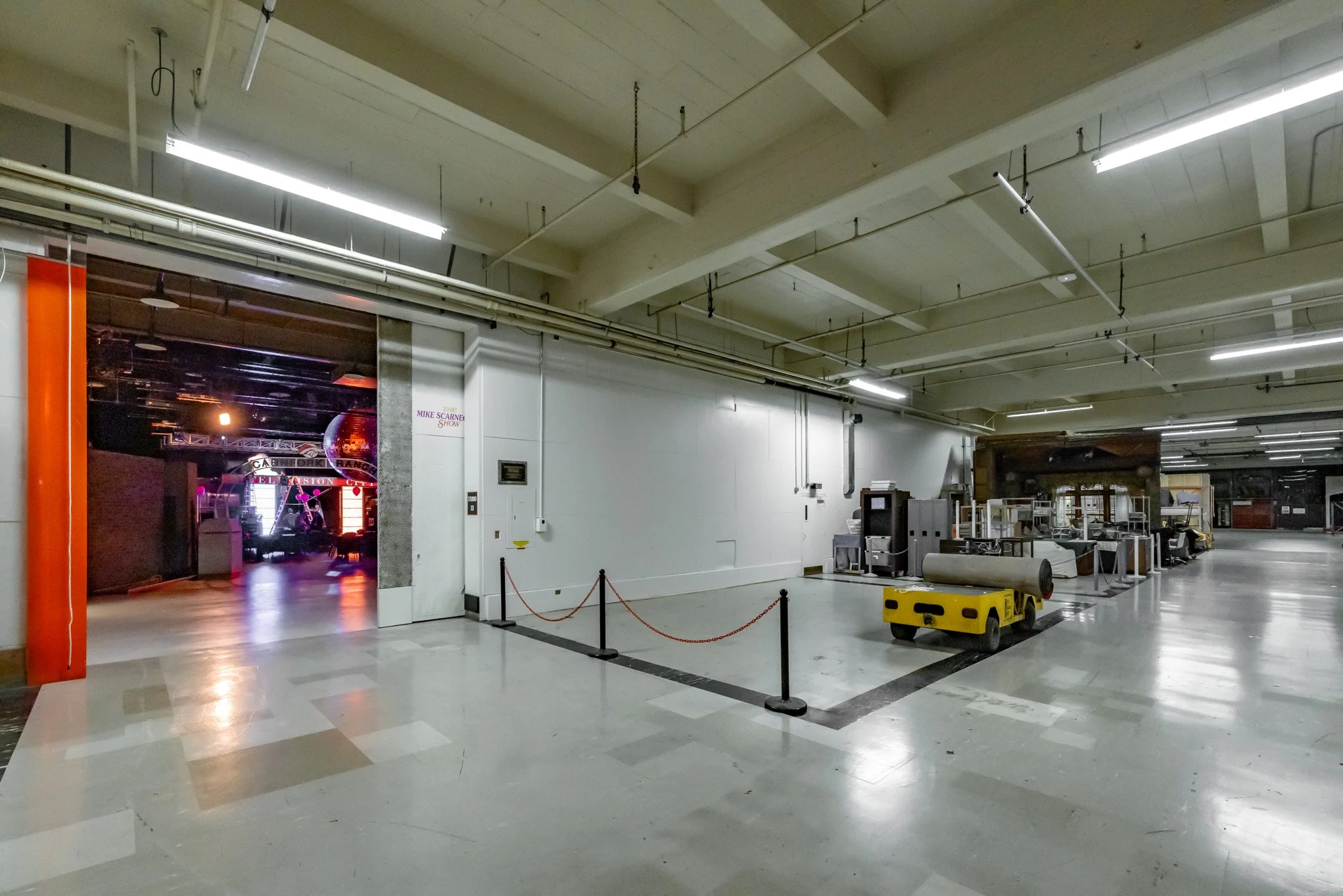Empty backstage area with industrial cleaning equipment and storage, doors to a neon-lit entertainment venue, and a spacious, unfinished look.