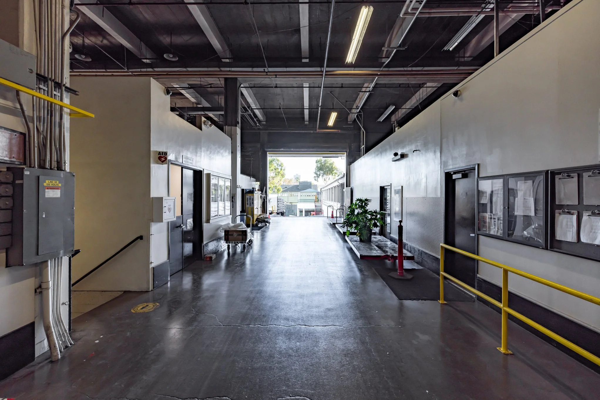 Empty hospital loading dock area with yellow safety railings, potted plants, and a large open gate at the end allowing sunlight and view of buildings outside.