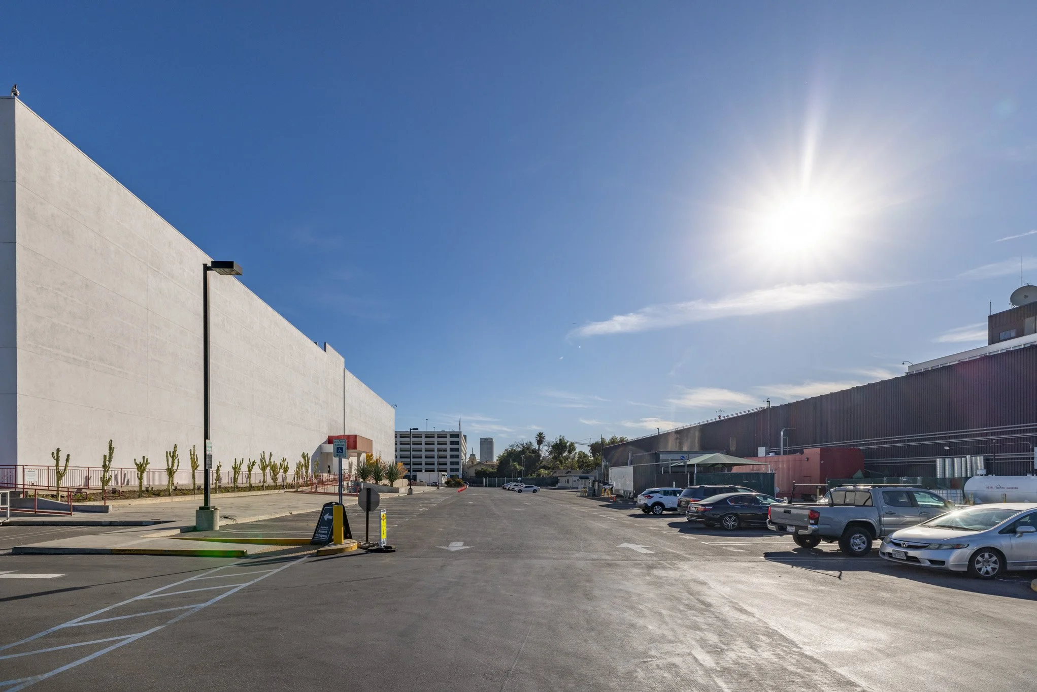 Empty parking lot in front of a large white building with cars parked on the side, under a bright sunny sky.