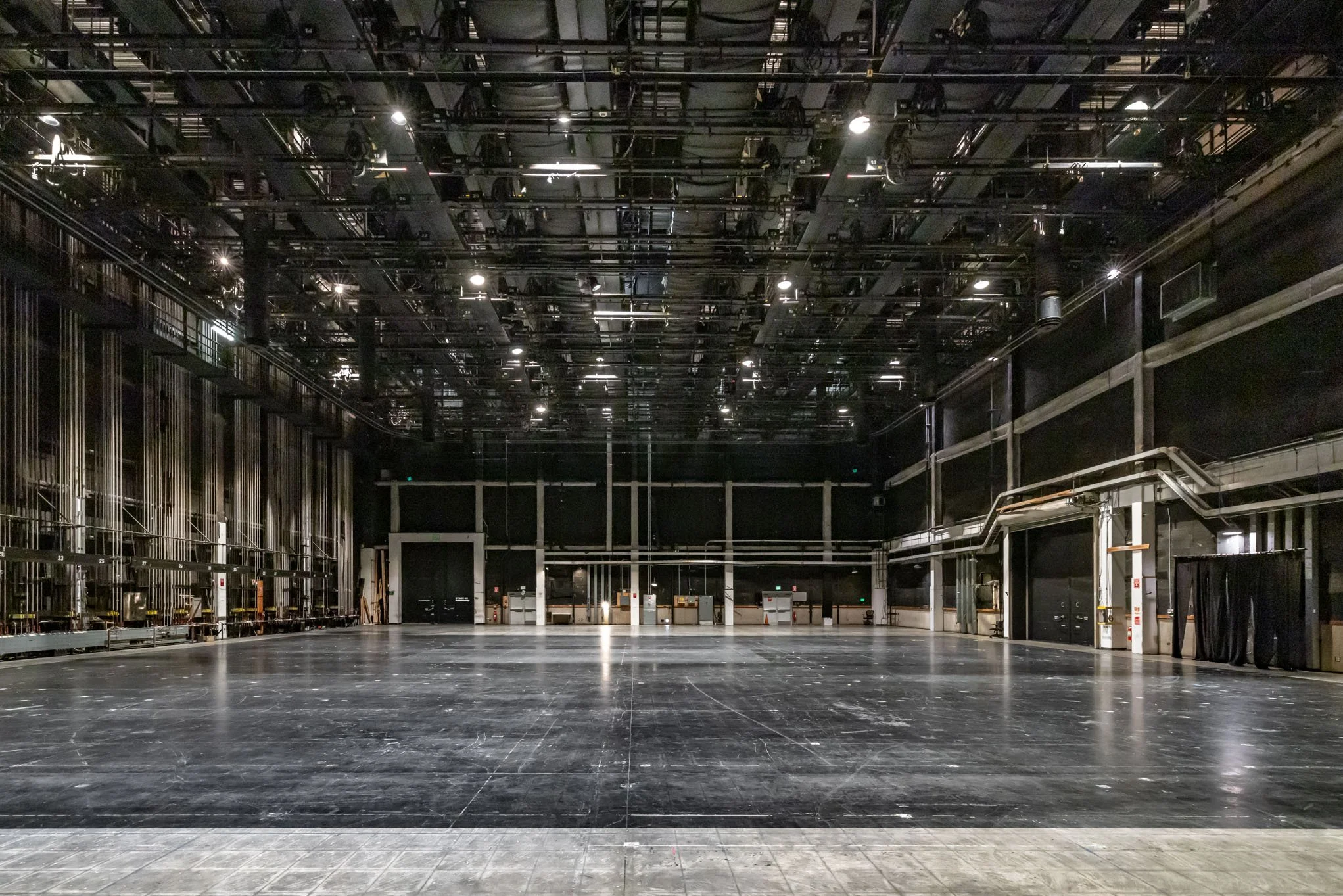 Empty stage or rehearsal space with black floor, black walls, and ceiling rigging, lights, and equipment.