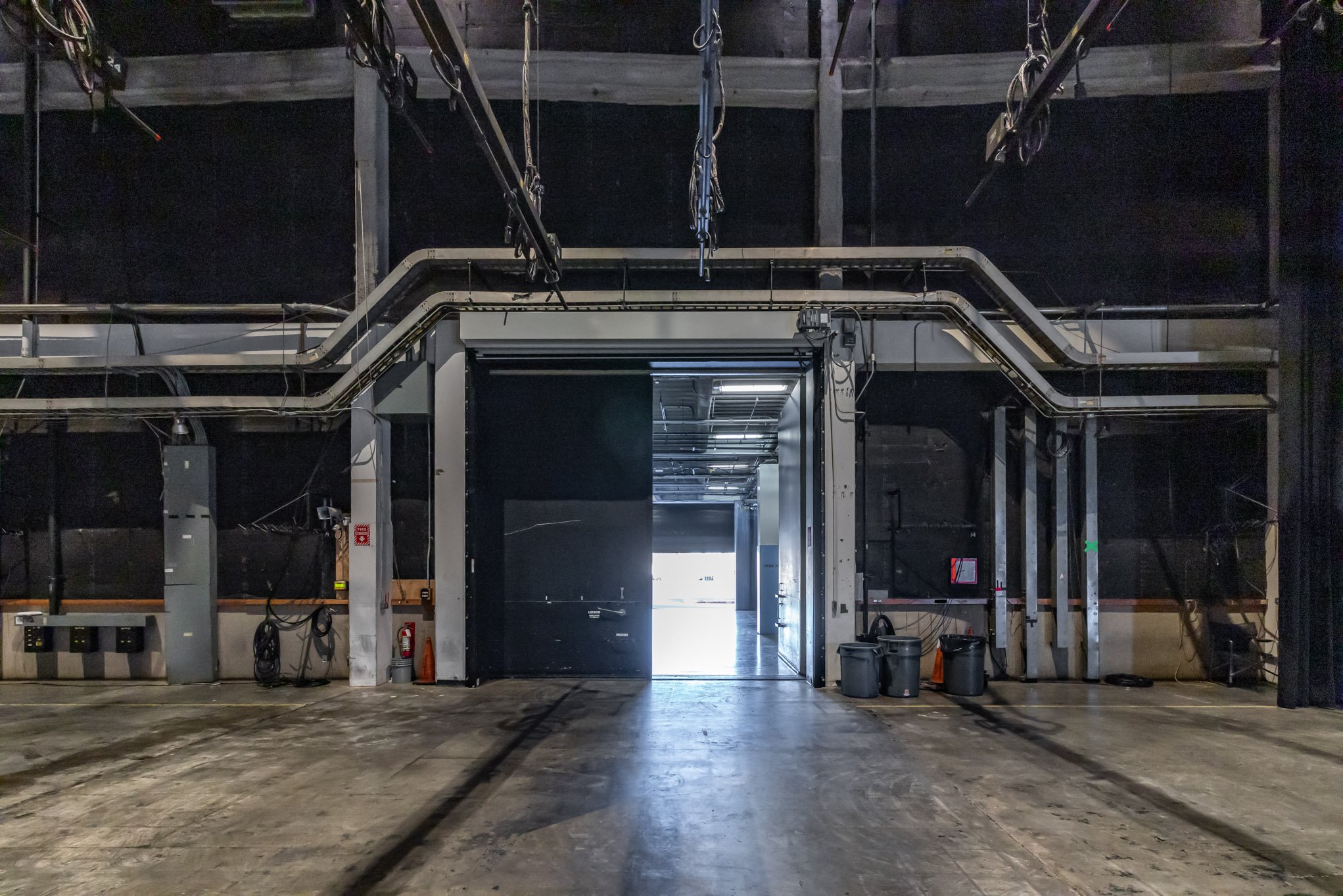 Empty event or warehouse space with industrial lighting, black walls, and a large open doorway illuminated by natural light.