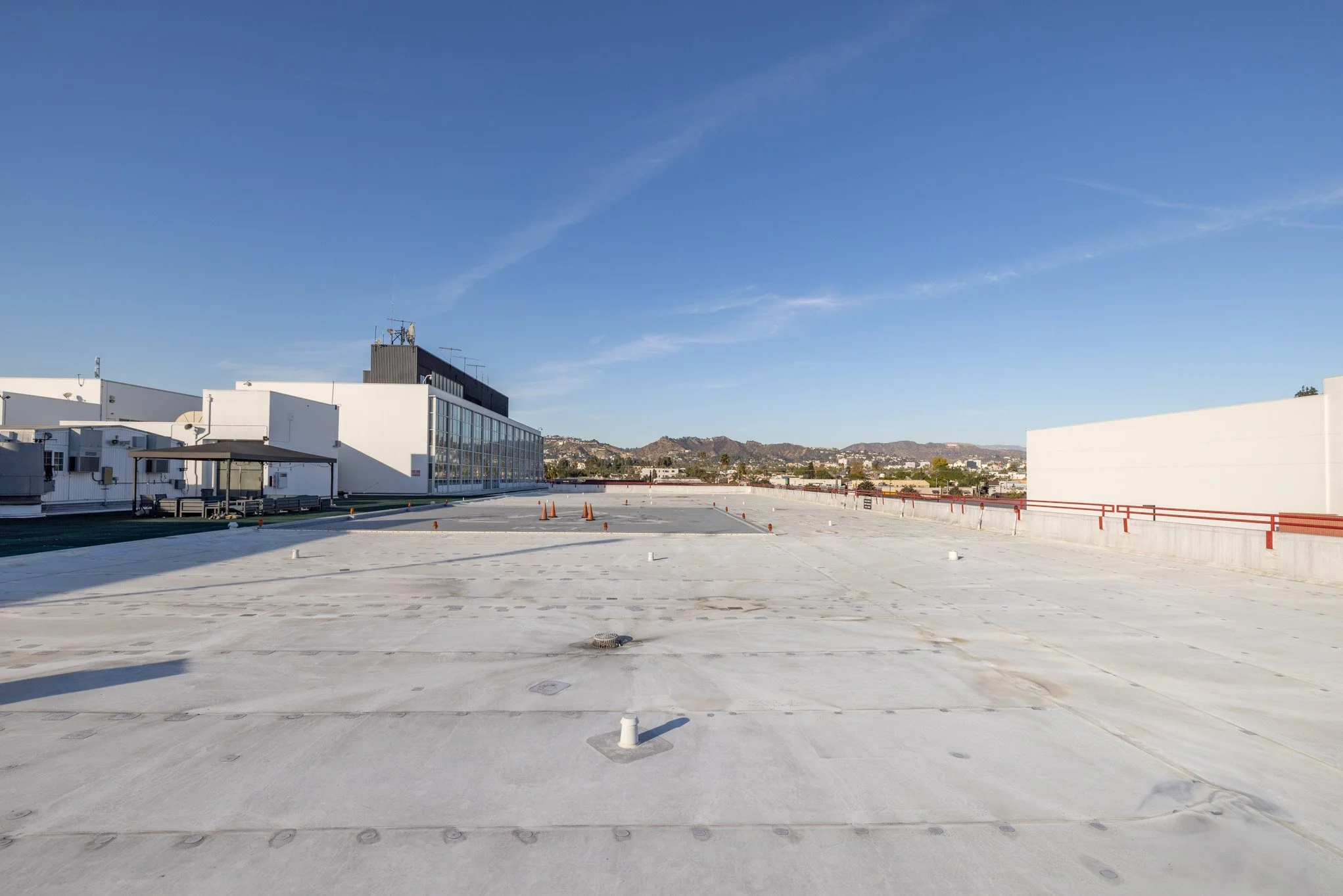 Empty rooftop parking area with safety cones and barriers, adjacent to a white commercial building with glass windows, with a cityscape and mountains in the background under a clear blue sky.