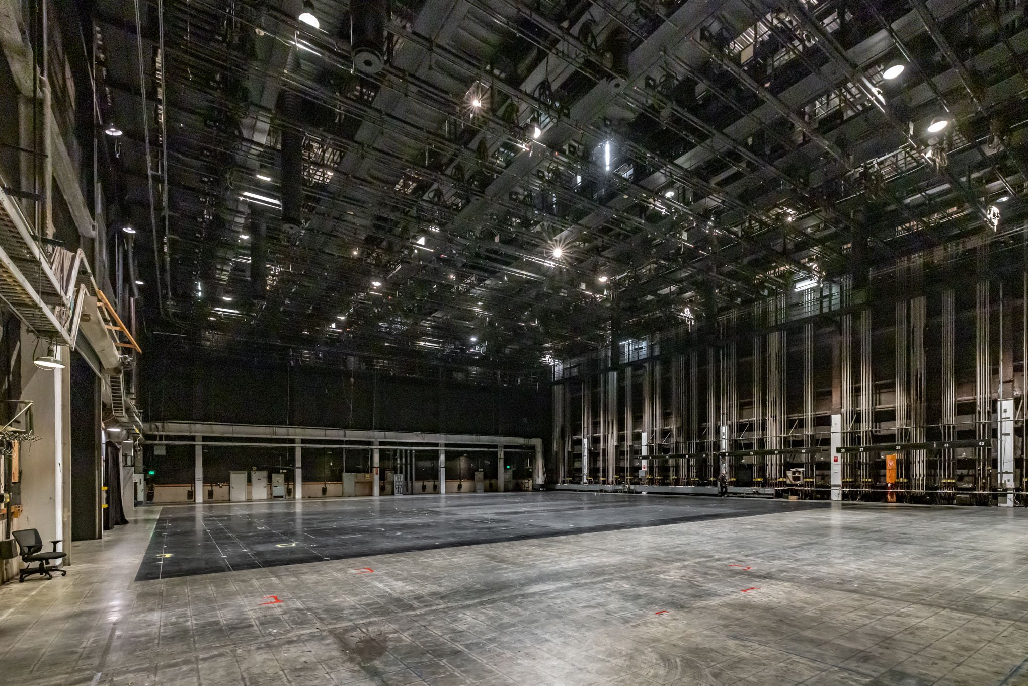 Empty stage in a theater with a black floor and a high, dark roof with stage lights and rigging.