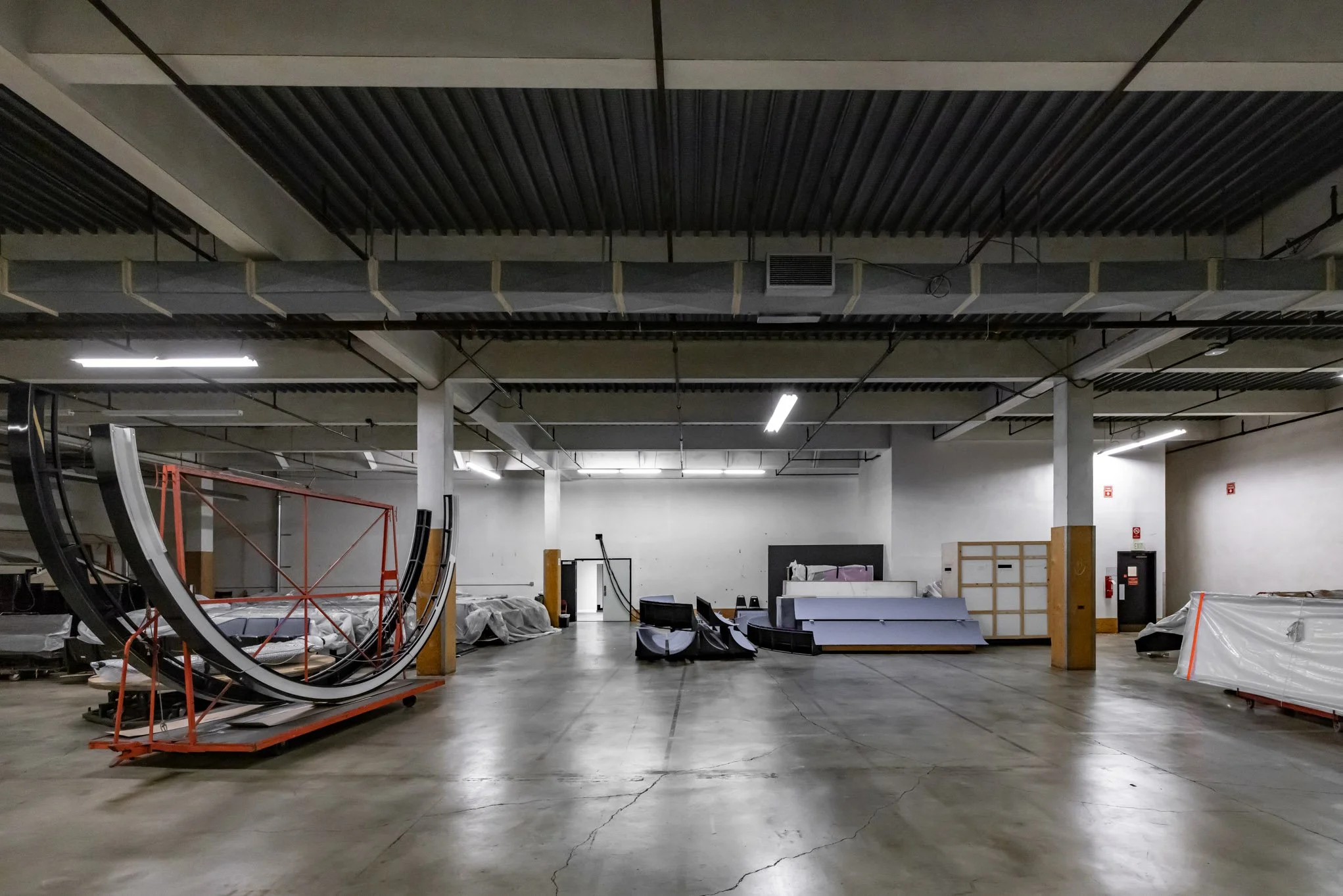 Empty indoor warehouse or storage room with construction or assembly materials, stacked on racks and covered in plastic, under fluorescent lighting.