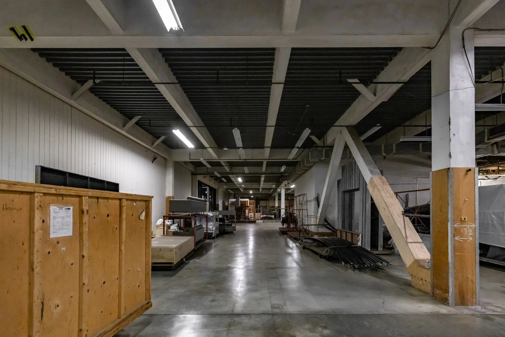 Empty retail store with scattered furniture, wooden crates, and exposed infrastructure, under fluorescent lighting.