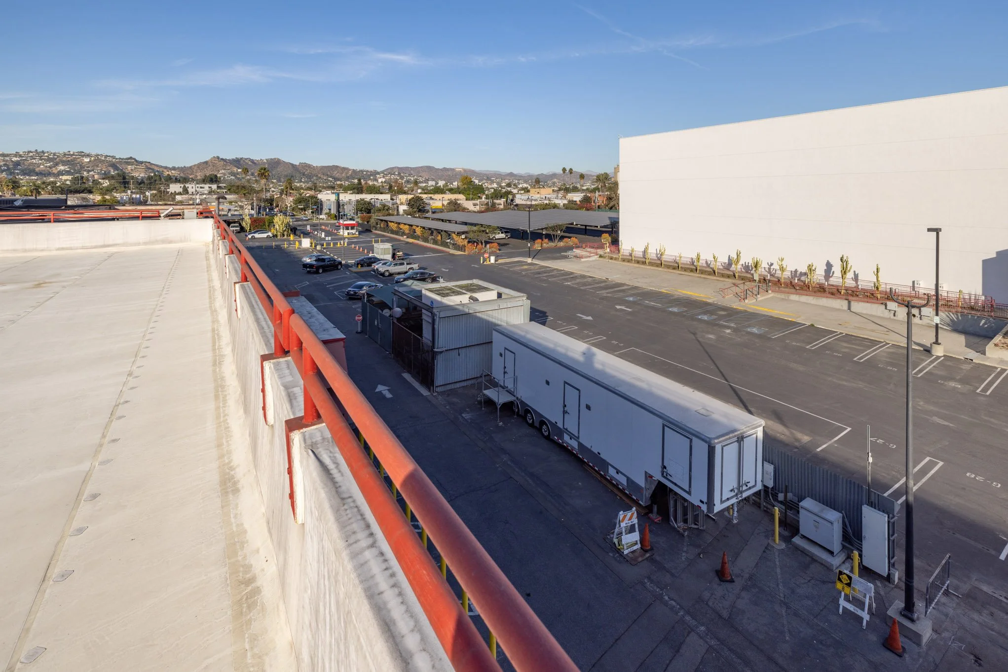 Empty parking lot with a truck and trailer, a white building, and a cityscape with hills in the background under a clear blue sky.