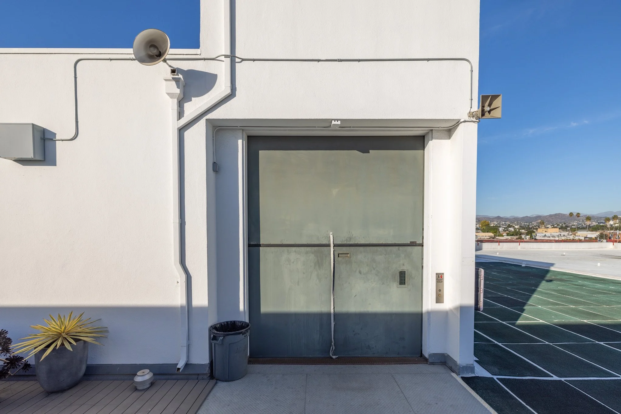 White building rooftop with a large gray elevator door, a trash can, a potted plant, and a stadium track with a cityscape and mountains in the background under a clear blue sky.