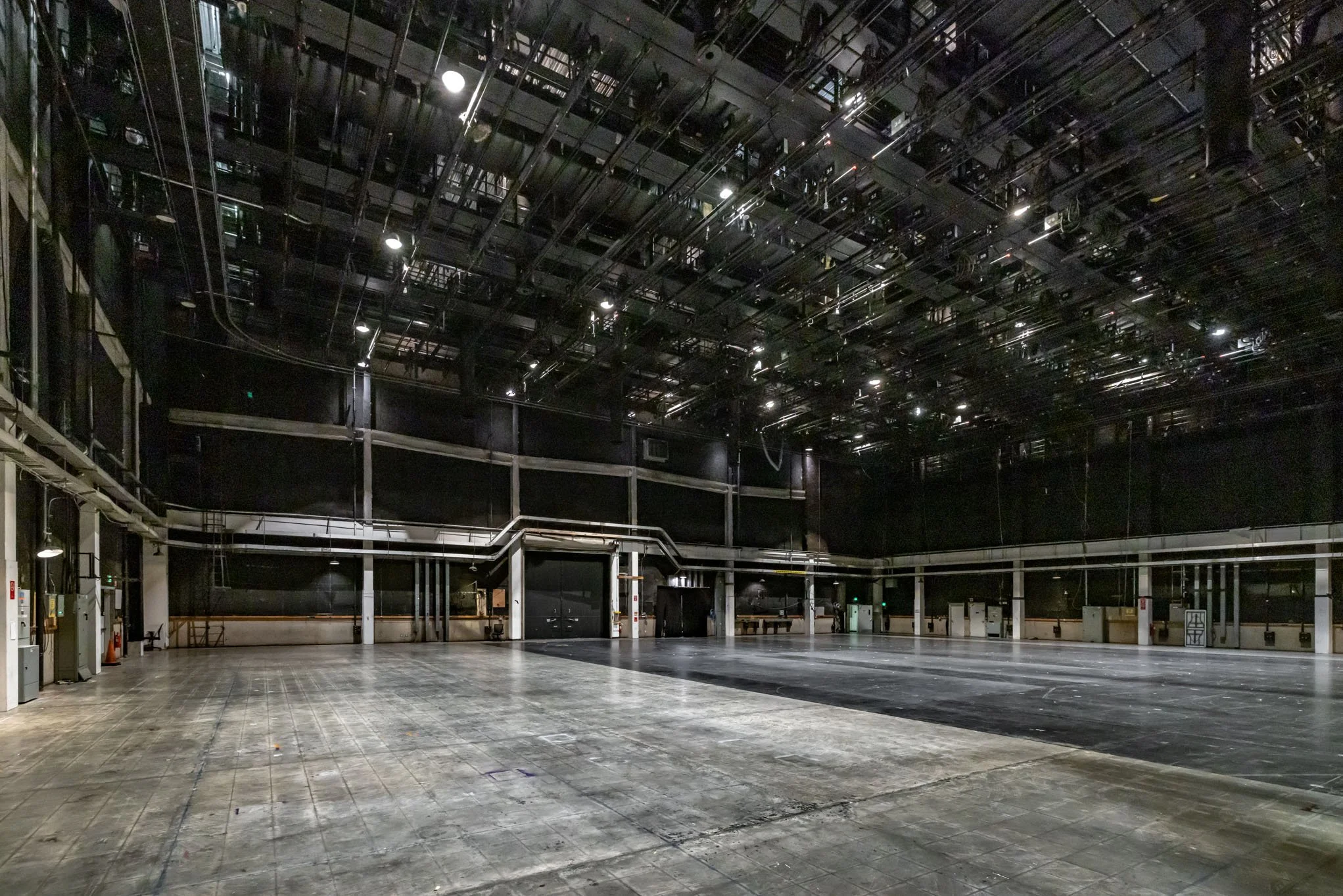 Empty stage or theater with stage floor, black walls, and overhead lighting equipment.