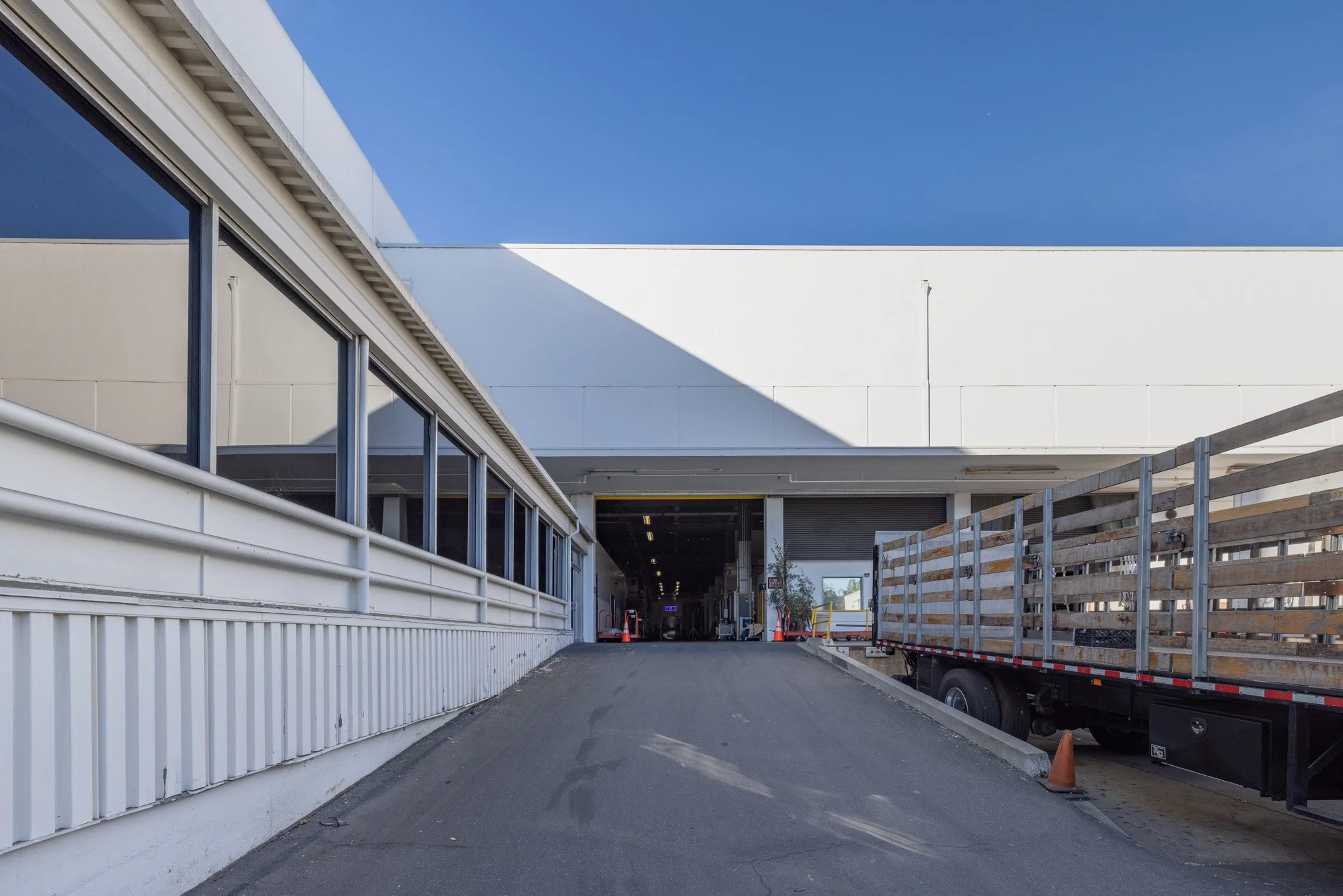 Loading dock entrance with a sloped driveway, parked delivery truck on the right, and a building with glass windows and a white facade. Orange cones are placed near the entrance.