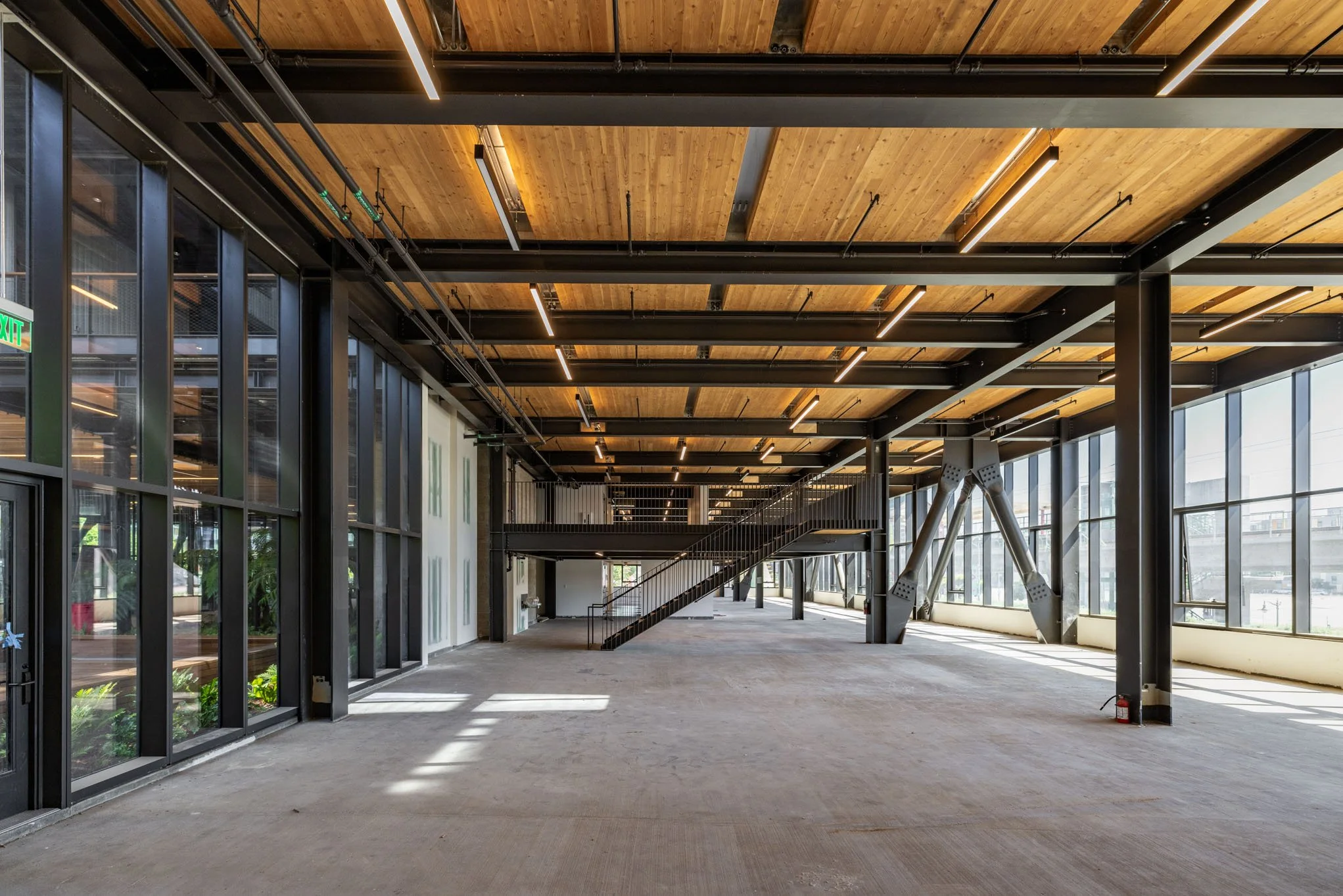 Modern, spacious interior of a building under construction with large glass windows, a wooden ceiling with black exposed beams, and a mezzanine level with stairs.