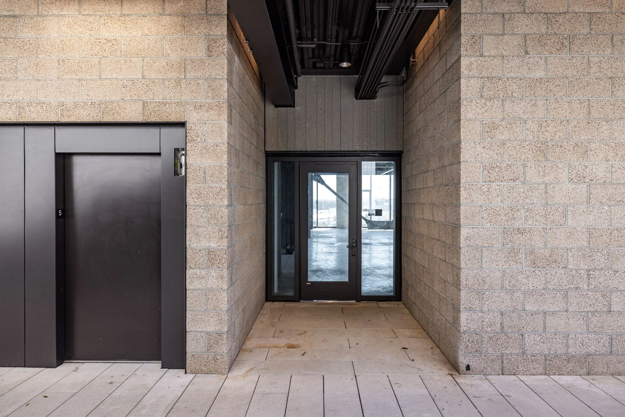 Inside view of an unfinished industrial building lobby with beige brick walls, a glass door, and concrete flooring with scattered debris.