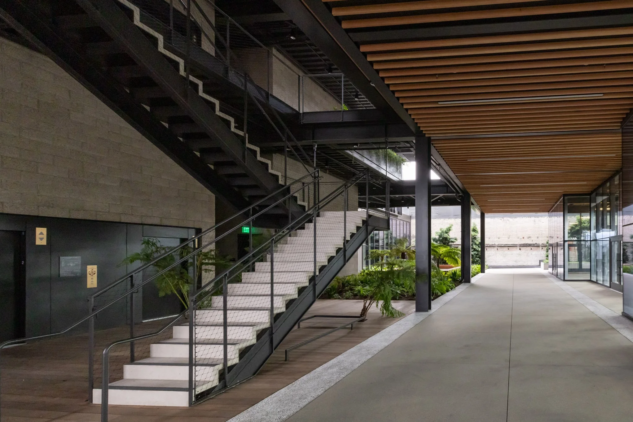 Modern indoor space with black metal staircase, glass walls, wooden ceiling, and indoor plants.