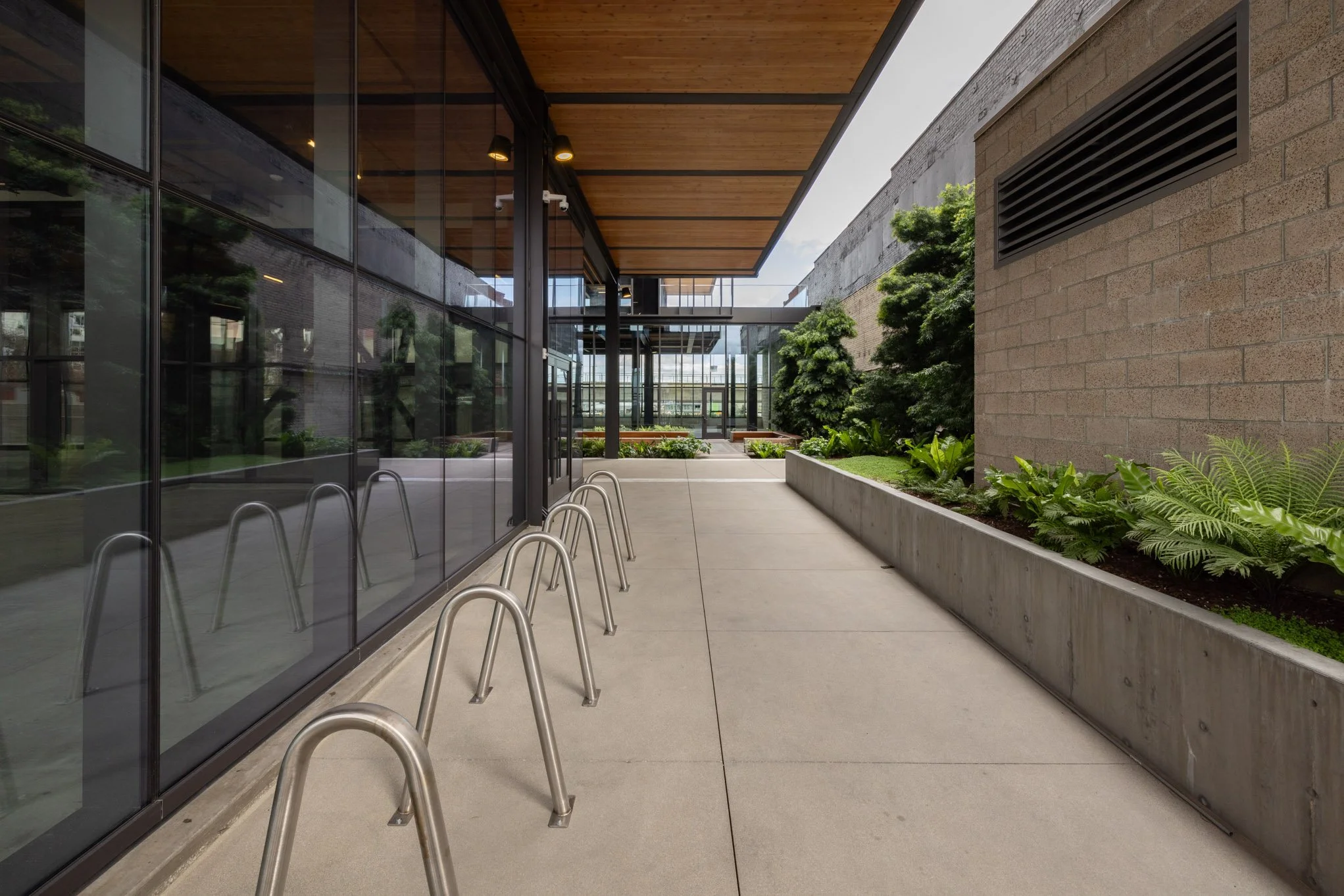 Modern building exterior with glass walls, metal bike racks, concrete sidewalk, and landscaped plants