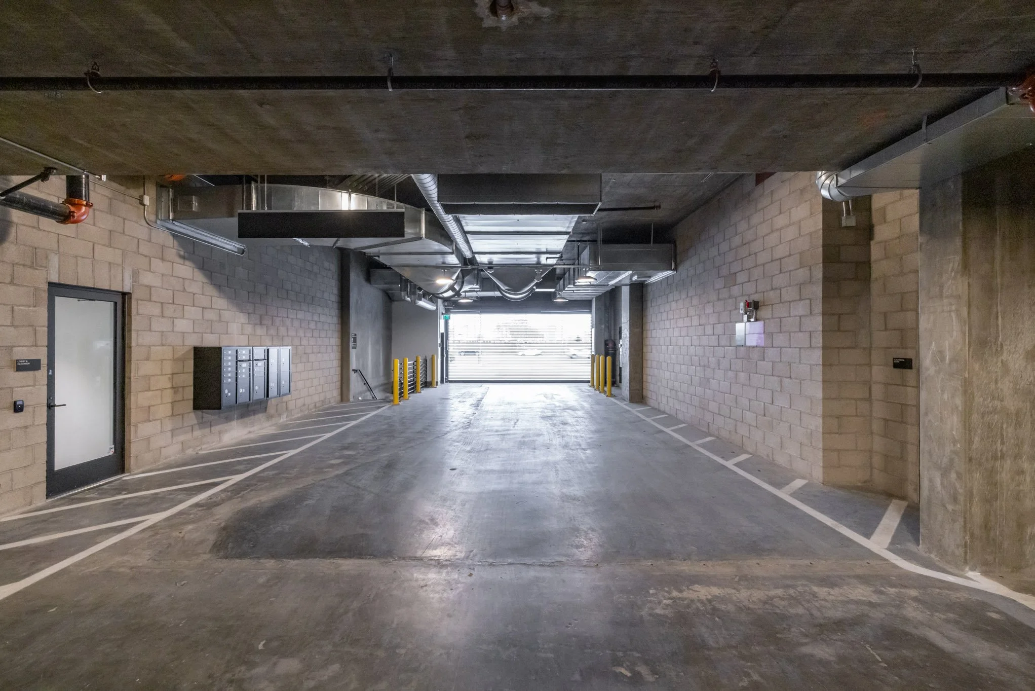 Underground parking garage with concrete floors and brick walls, illuminated by natural light from open entrance, featuring yellow posts, black mailboxes, and a door on the left side.
