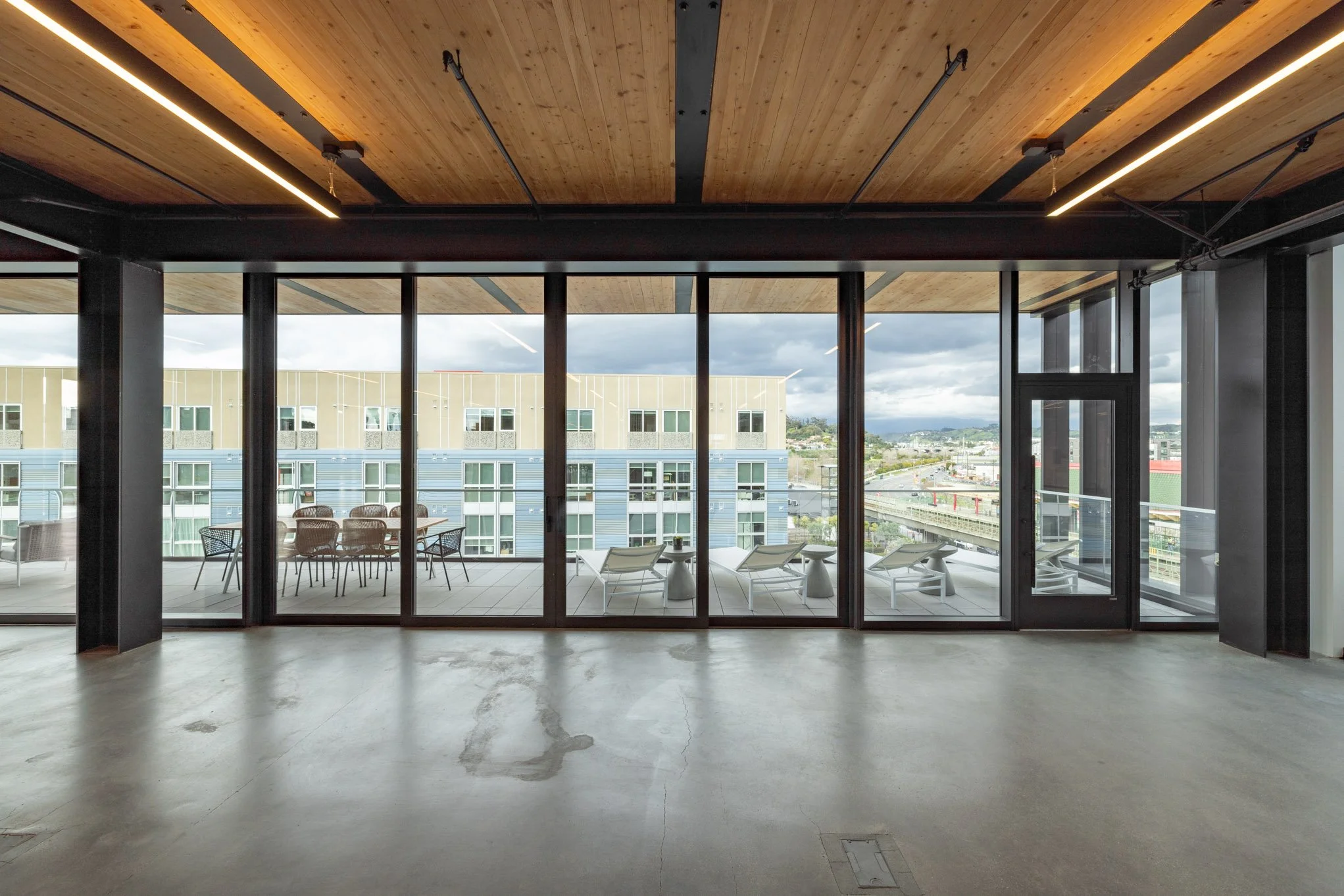 Empty room with large glass sliding doors leading to a balcony with outdoor chairs and tables, overlooking a cityscape with modern buildings and cloudy sky.