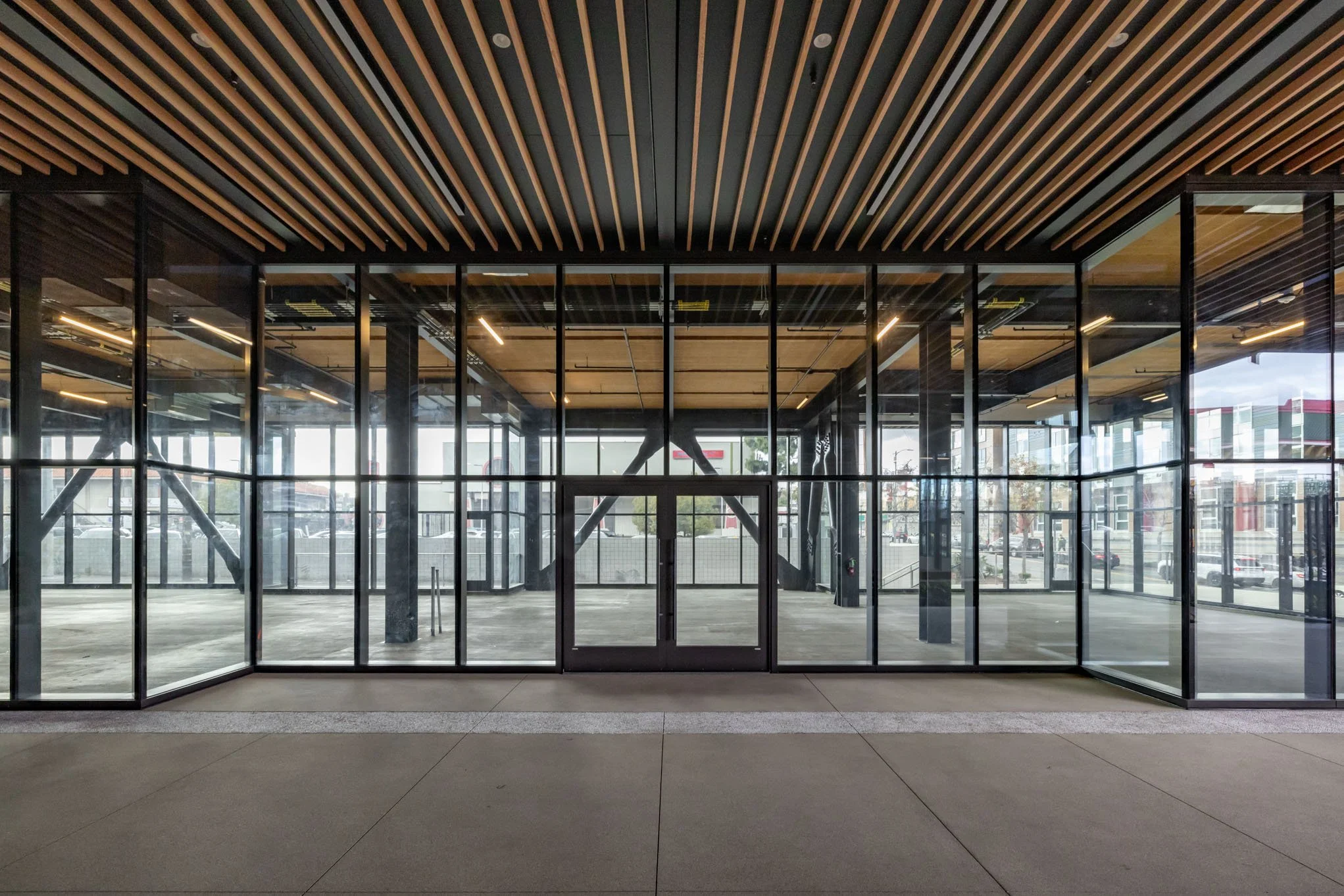Modern building entrance with glass walls and wooden ceiling panels, view inside toward an open space with structural supports, and parking lot visible outside.