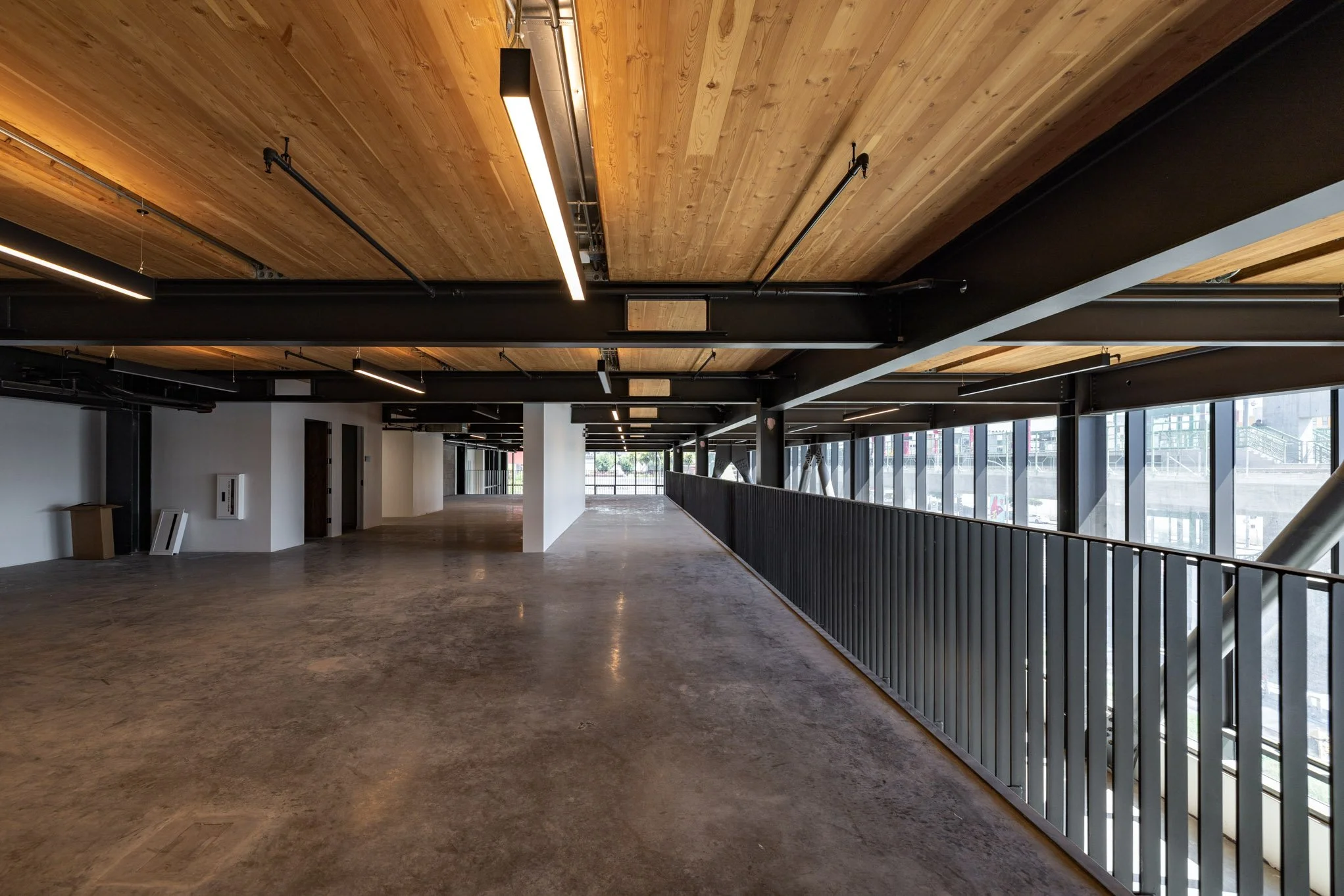 Empty interior space with concrete floor, black steel beams, wood-paneled ceiling with modern lighting, large windows, and a metal railing along a walkway.