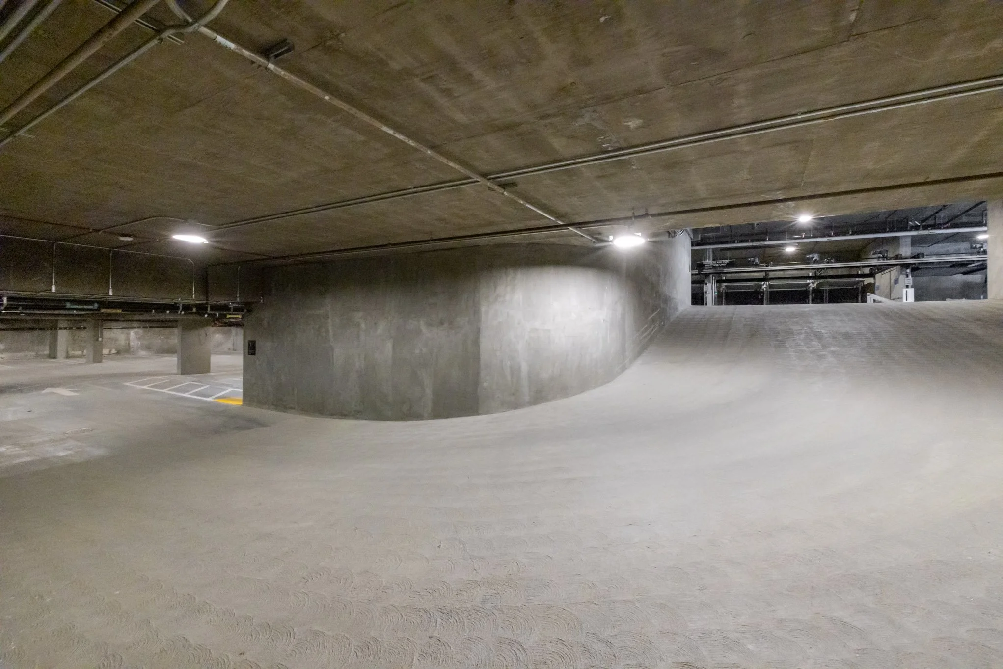 Underground parking garage with concrete walls, ceiling, and floor, illuminated by ceiling lights.