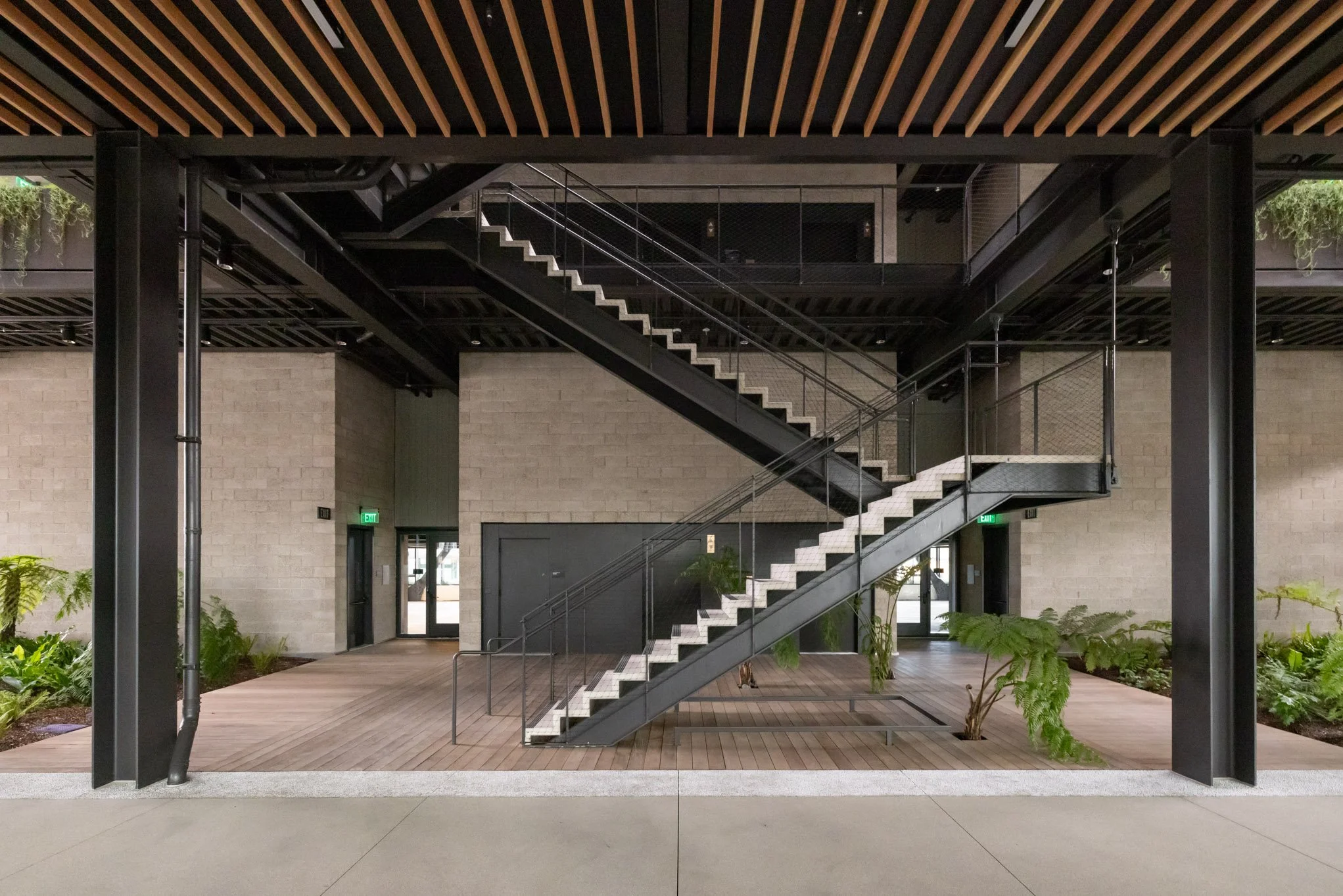 Interior view of a modern building with black metal staircase, wooden flooring, beige brick walls, and green plants.