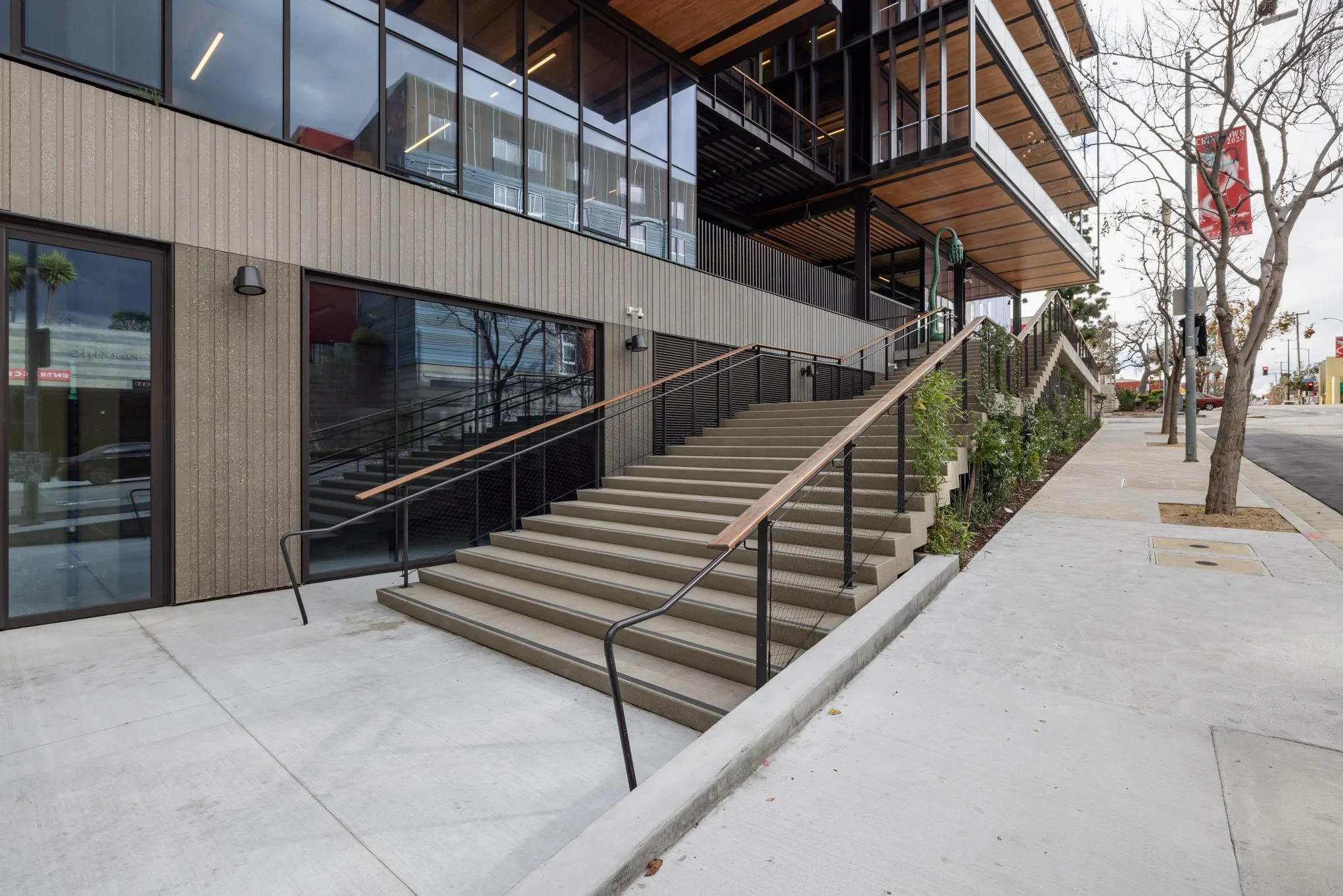Exterior of a modern building with concrete stairs, metal railings, glass windows, and a sidewalk with trees.