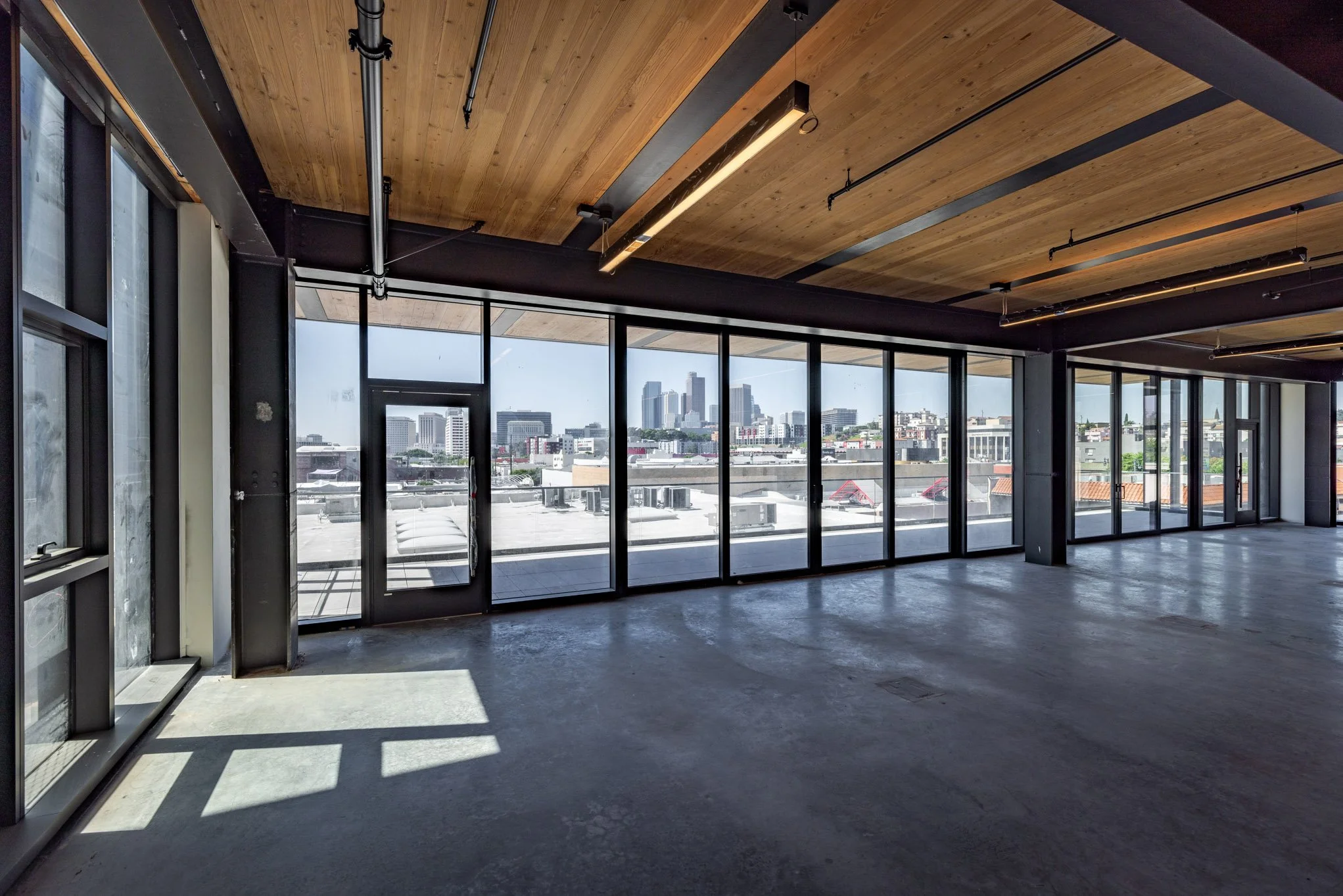 Empty commercial space with large floor-to-ceiling windows, a concrete floor, wooden ceiling, and city skyline view.